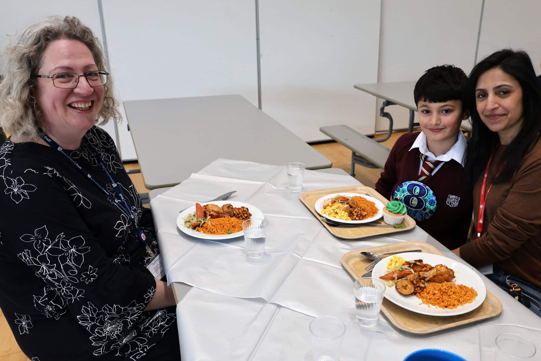 Three people, two adults and a child, sit at a canteen table with plates of food, smiling at the camera. The table is covered with a white cloth and glasses of water are present.