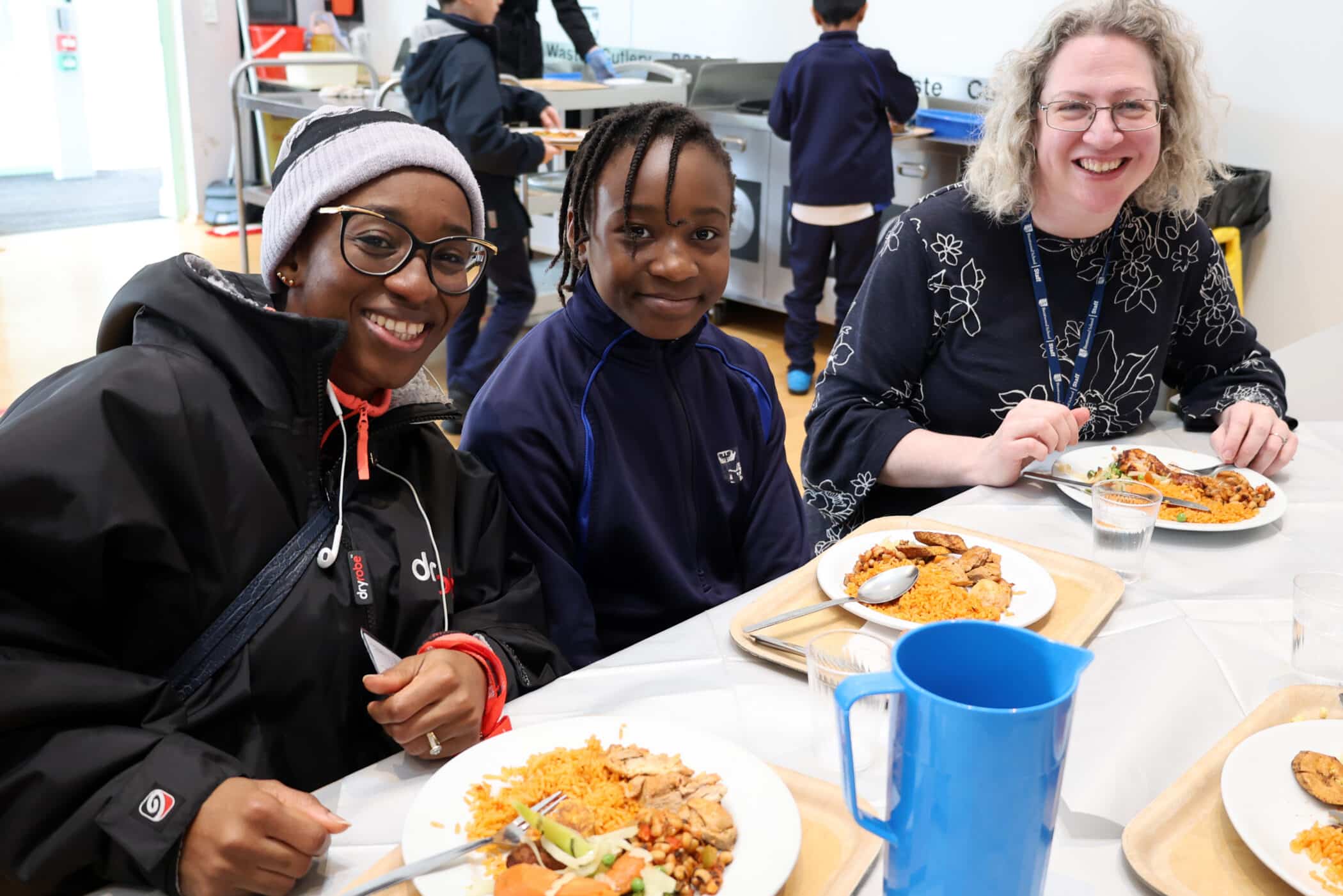Three people are sitting at a table and smiling while eating a meal that includes rice, chicken, and salad. There are trays, plates, and a blue water jug on the table. Other people are visible in the background.