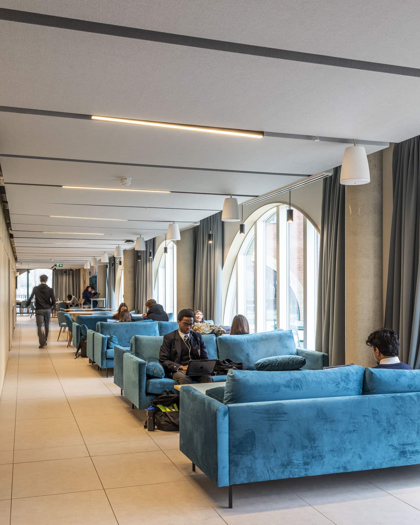 People sit on bright blue sofas in a modern lounge with large arched windows and light floors, chatting about topics like day school vs boarding school, while others work on laptops or walk through the space.