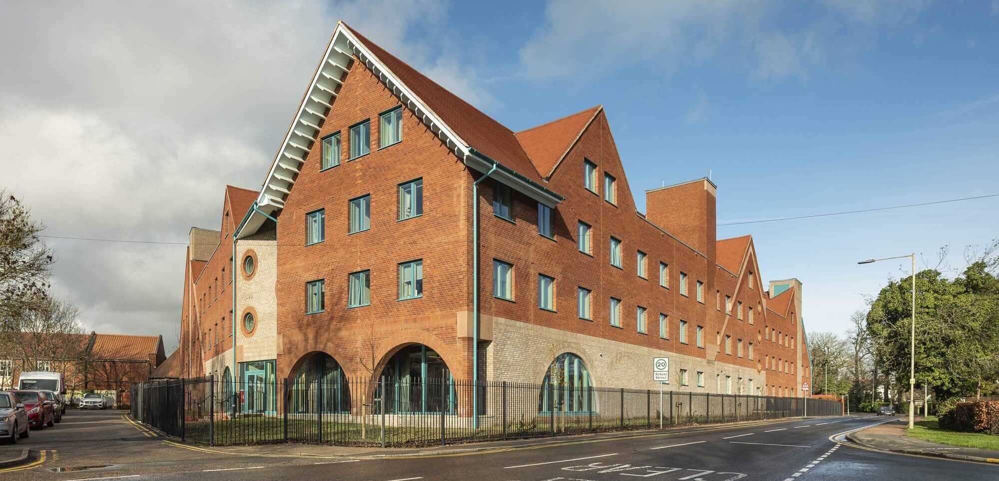A large, modern red-brick building with steep, pointed roofs and blue window frames stands at a street corner under a partly cloudy sky, an impressive sight that suits a boarding school behind its black fence and arched entrances.