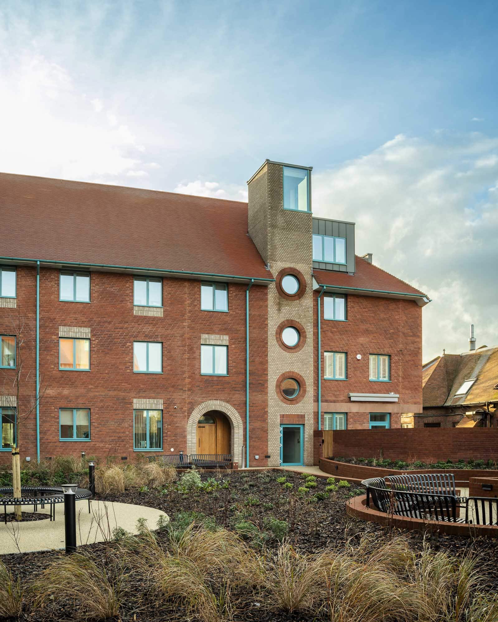A modern brick boarding building with a red sloping roof, tall glass tower, and landscaped gardens mirrors how boarding schools develop independence, offering spaces for students to grow amidst pathways and benches under a partly cloudy sky.
