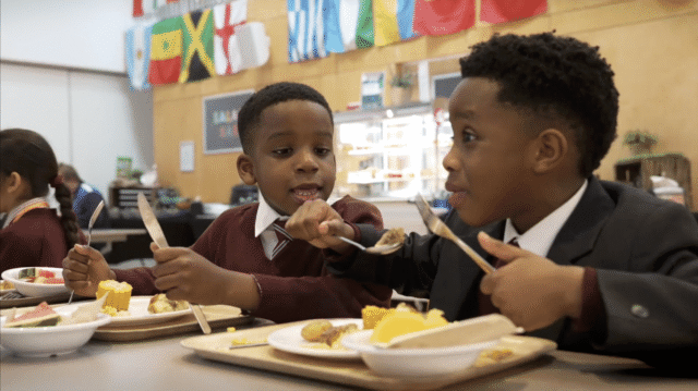 Two boys in school uniforms sit at a canteen table, eating lunch and smiling at each other. Colourful international flags hang above them, and food trays with various items are on the table.