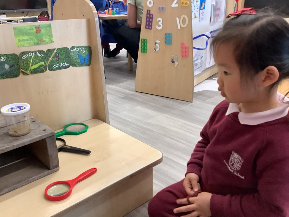 A young girl in a burgundy school uniform observes a small container with what appears to be a caterpillar inside, using magnifying glasses on a table in a classroom setting with educational displays.