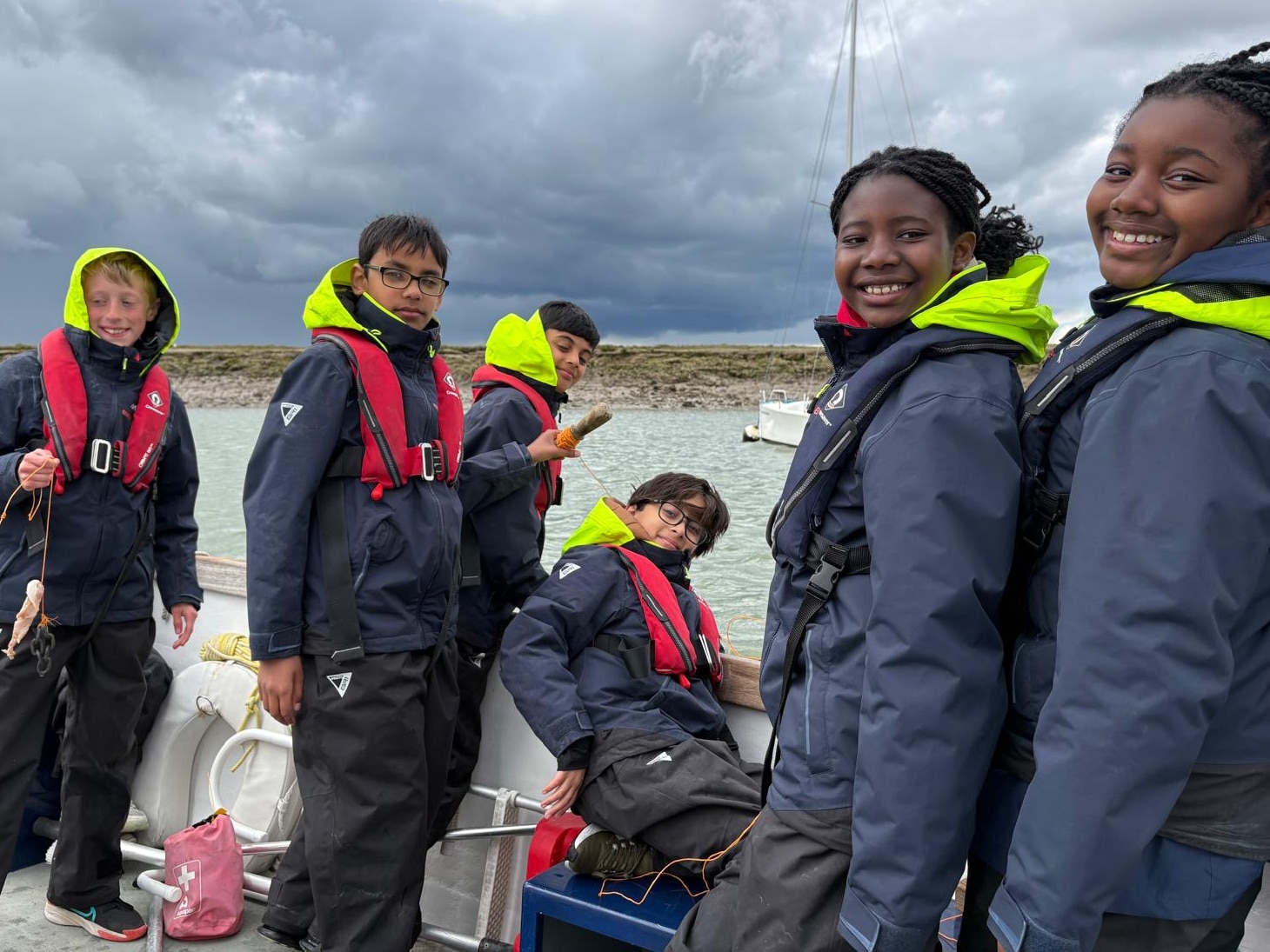 Six children wearing navy waterproof jackets with neon hoods and red lifejackets stand and sit on a boat, smiling. The sky is cloudy, and a yacht and shoreline are visible in the background.
