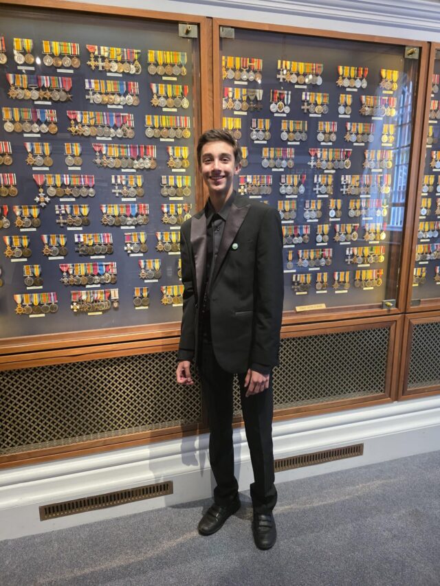 A young man in a black suit stands smiling in front of a large display case filled with rows of colourful military medals and ribbons in a museum or exhibition setting.