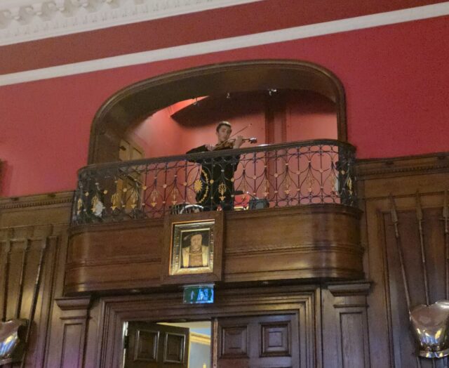 A person in uniform stands on an upper balcony in a grand hall with red walls, wooden panelling, and large framed portraits of people in regal attire hanging on either side.