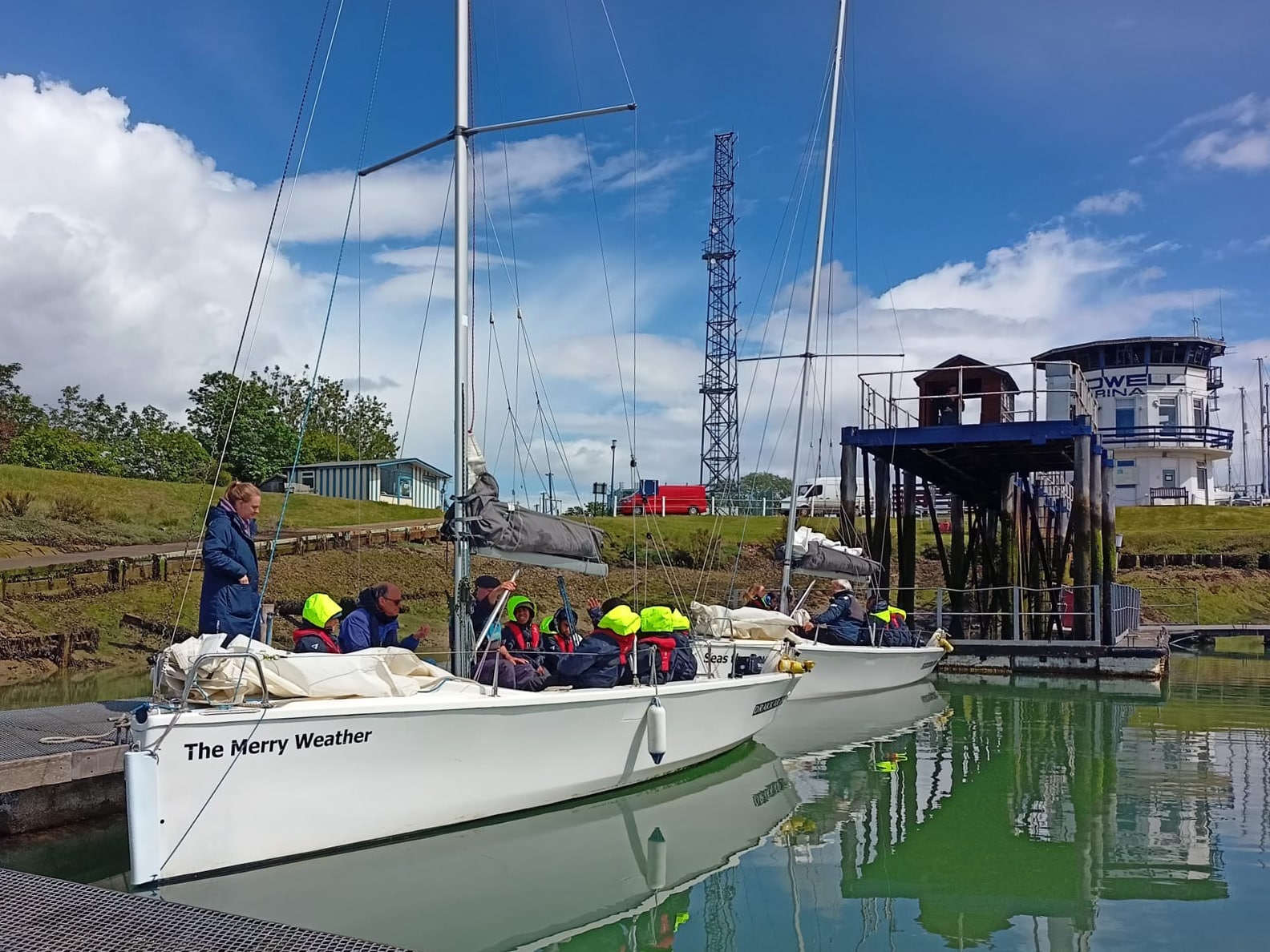 Two small sailing boats named The Merry Weather are moored in calm water. Several people wearing bright yellow life jackets sit on board, while a few stand nearby. A blue sky with clouds and various buildings are in the background.