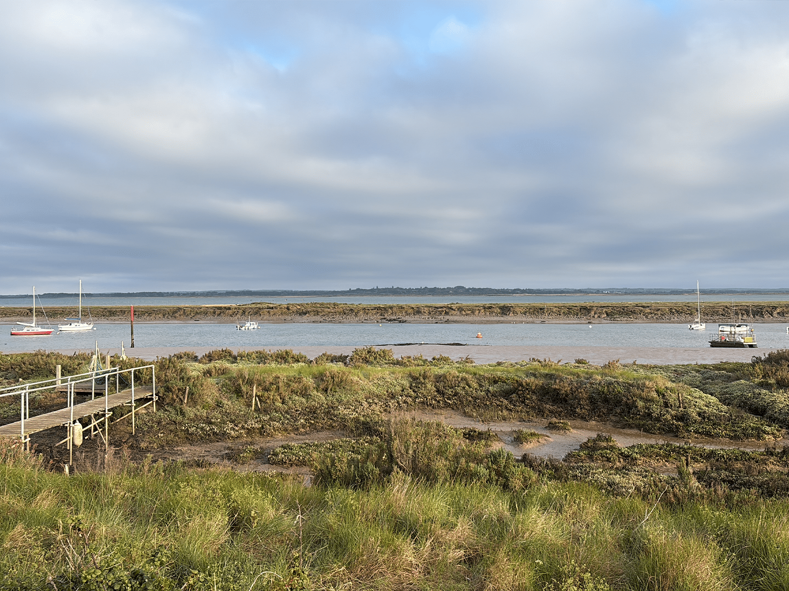 A calm river bordered by grassy marshland under a cloudy sky, with several small sailing boats anchored on the water and a wooden jetty on the left side of the image.
