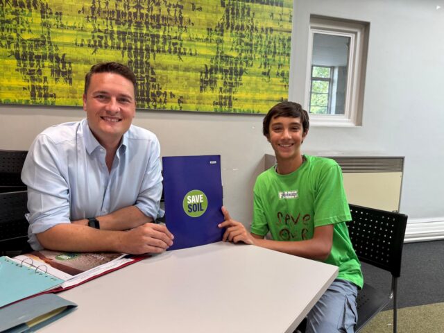 Two people sitting at a table indoors, smiling at the camera. One holds a folder that says SAVE SOIL, whilst the other wears a green Save Soil T-shirt. A modern, abstract painting hangs on the wall behind them.
