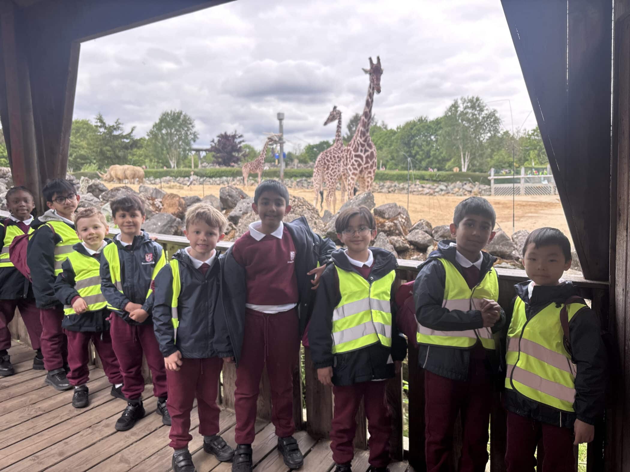 A group of schoolchildren in maroon uniforms and yellow reflective vests stand smiling on a wooden platform at a zoo, with giraffes and other animals visible in the background.