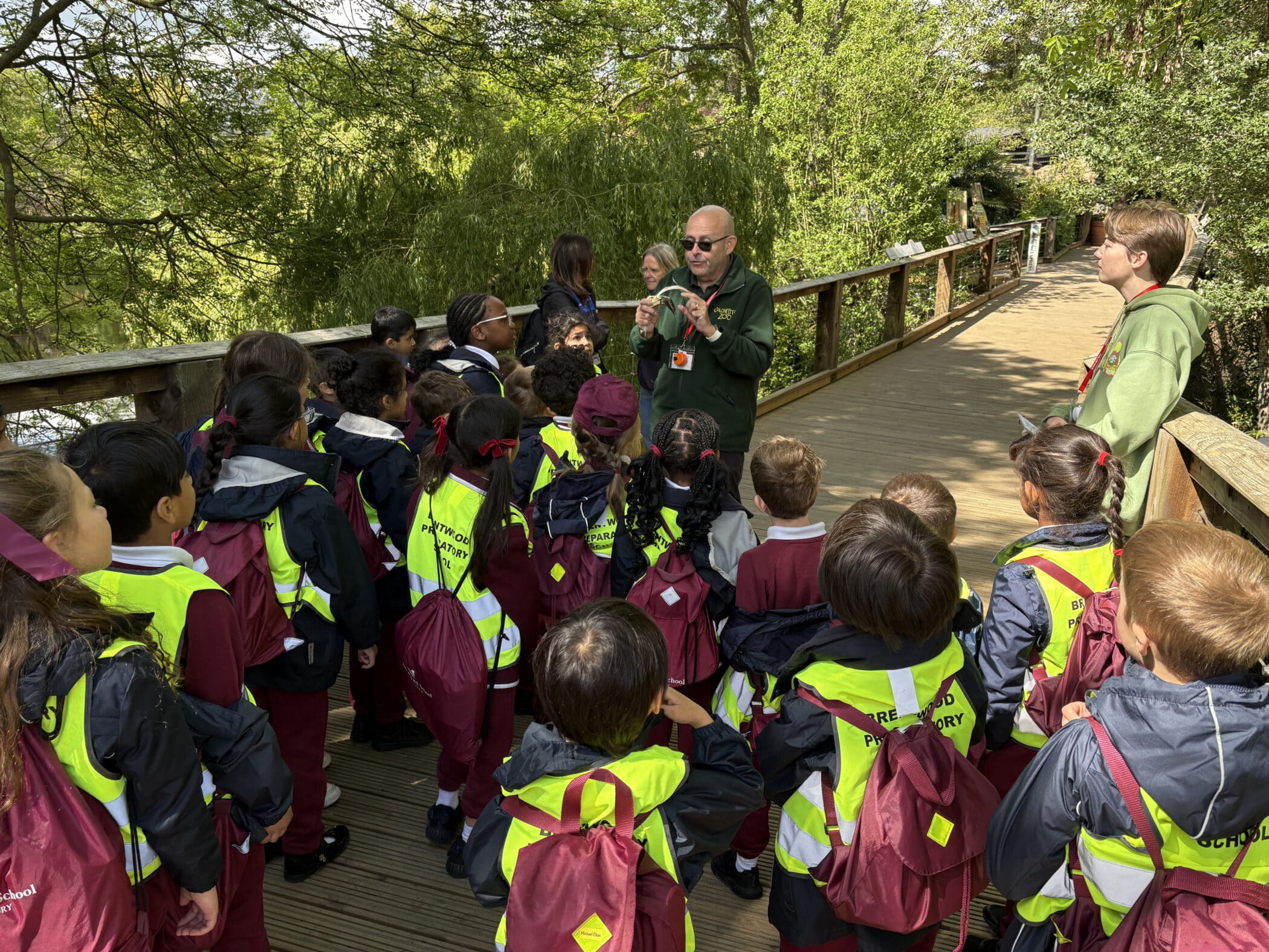 A group of young students wearing maroon uniforms and yellow safety vests listen to a guide speaking on a wooden walkway surrounded by trees during an outdoor educational trip.