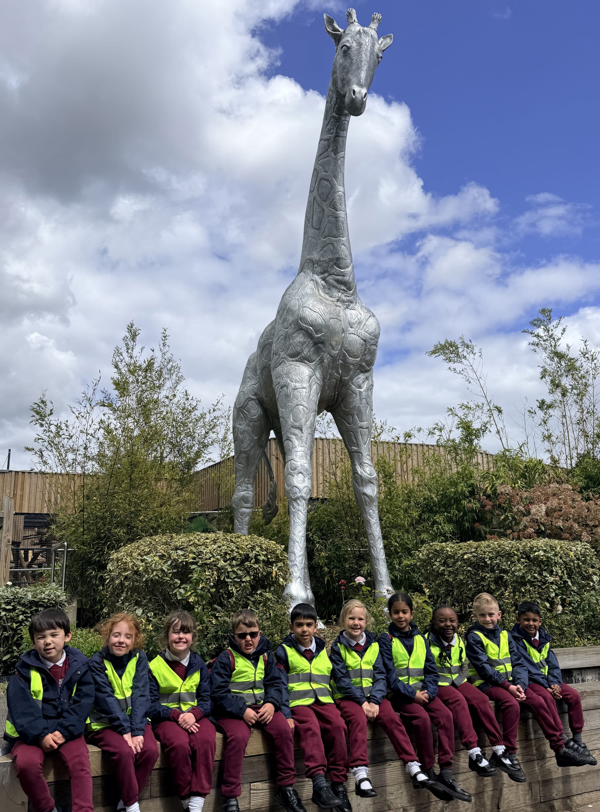 A group of young children in yellow safety vests and red uniforms sit in a row in front of a large metallic giraffe sculpture, with greenery and a cloudy blue sky in the background.