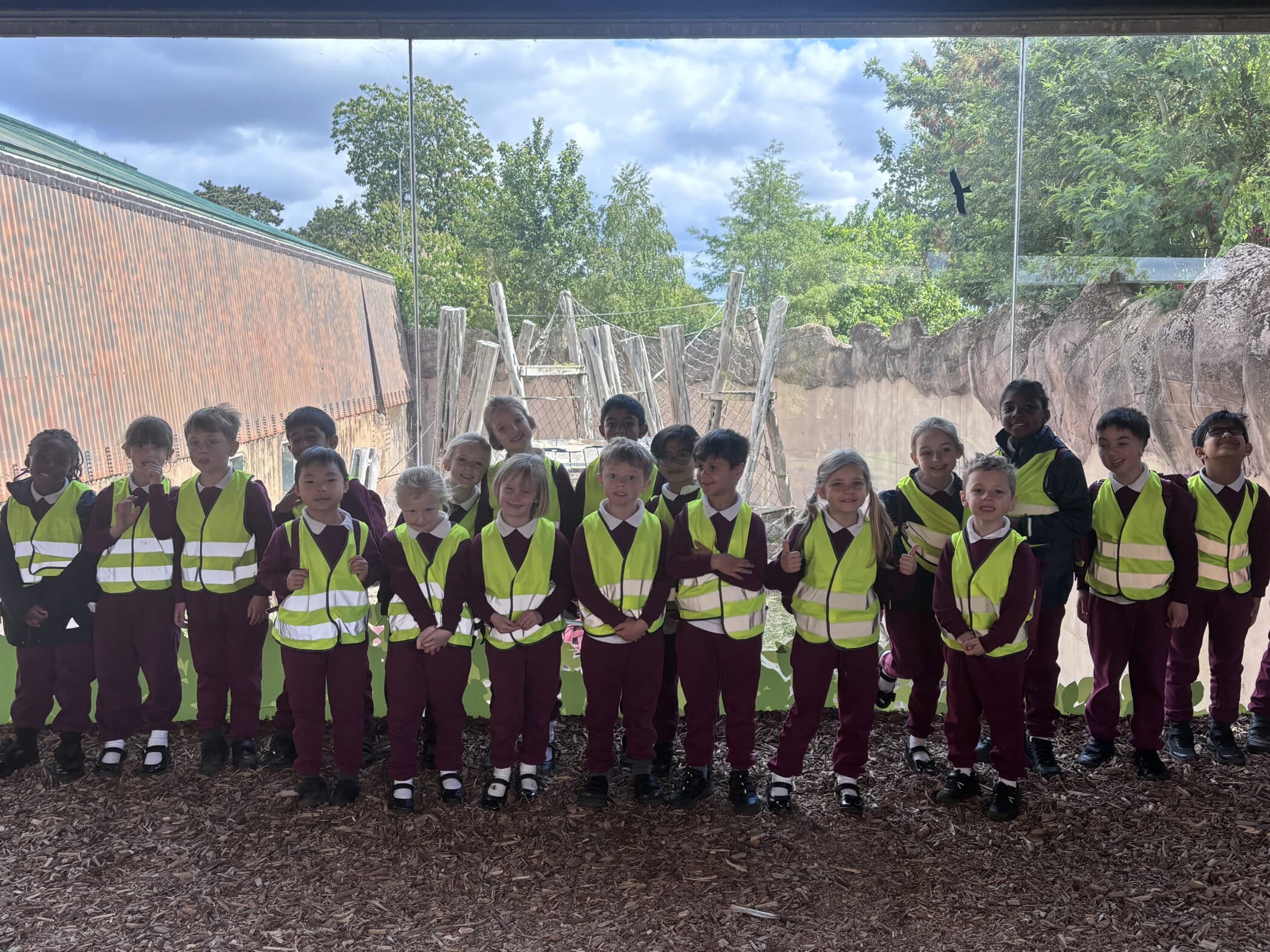 A group of young children wearing matching maroon outfits and reflective yellow vests stand together indoors in front of a large window with an outdoor play area and trees visible behind them.