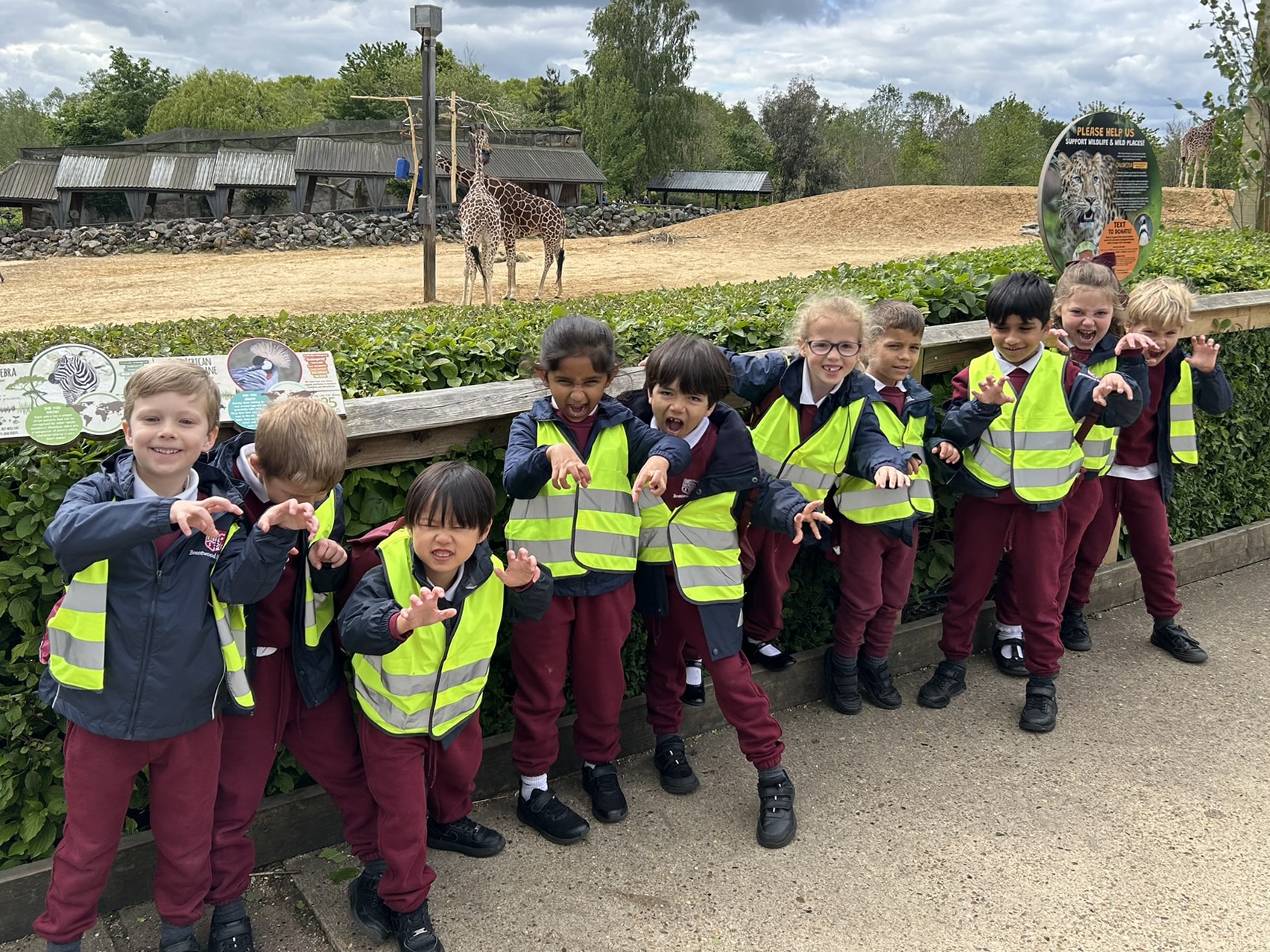 A group of smiling young children in maroon uniforms and yellow safety vests pose playfully in front of a giraffe enclosure at a zoo. Lush greenery and animal signs are visible in the background.