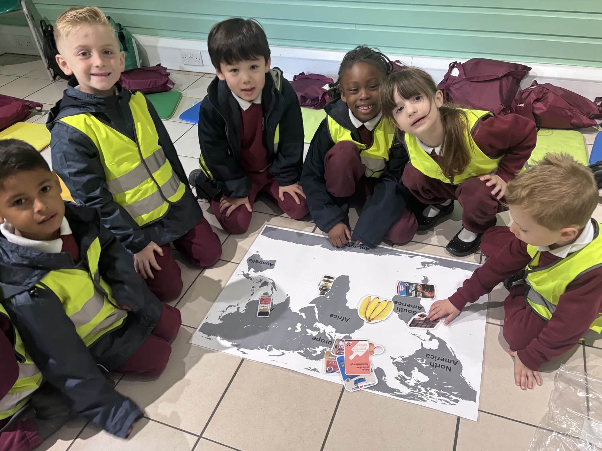 Six young children in yellow safety vests and school uniforms sit around a large world map on the floor, engaging with cards and objects placed on different continents. They are smiling and looking at the camera.