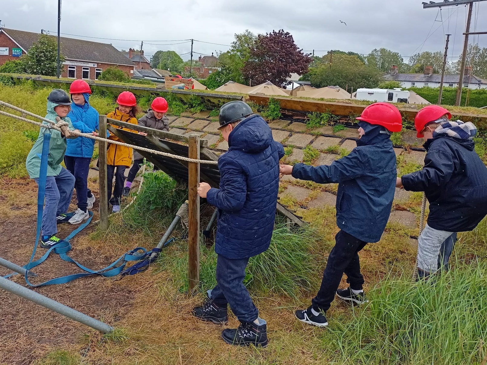 A group of children wearing red and black helmets work together outdoors to lift or adjust a wooden and rope structure on a grassy area, with buildings and trees visible in the background.