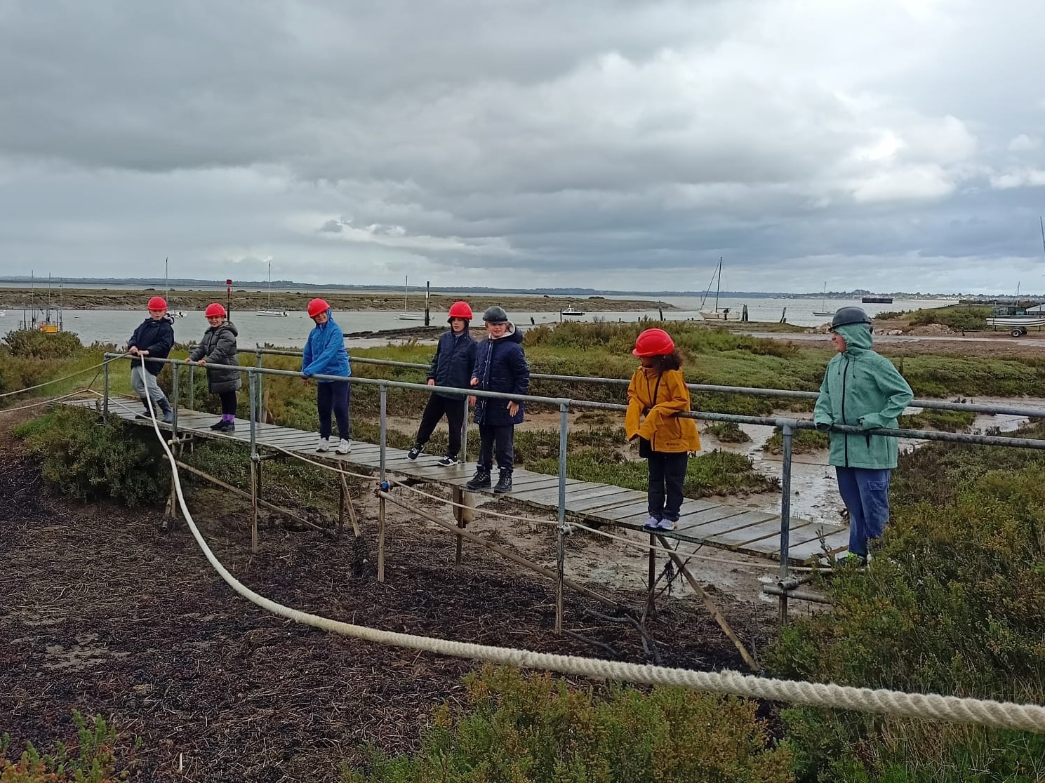 A group of people wearing red helmets and outdoor jackets stand on a narrow wooden footbridge and nearby path in a marshy area under a cloudy sky. Some are watching, while others cross the footbridge.