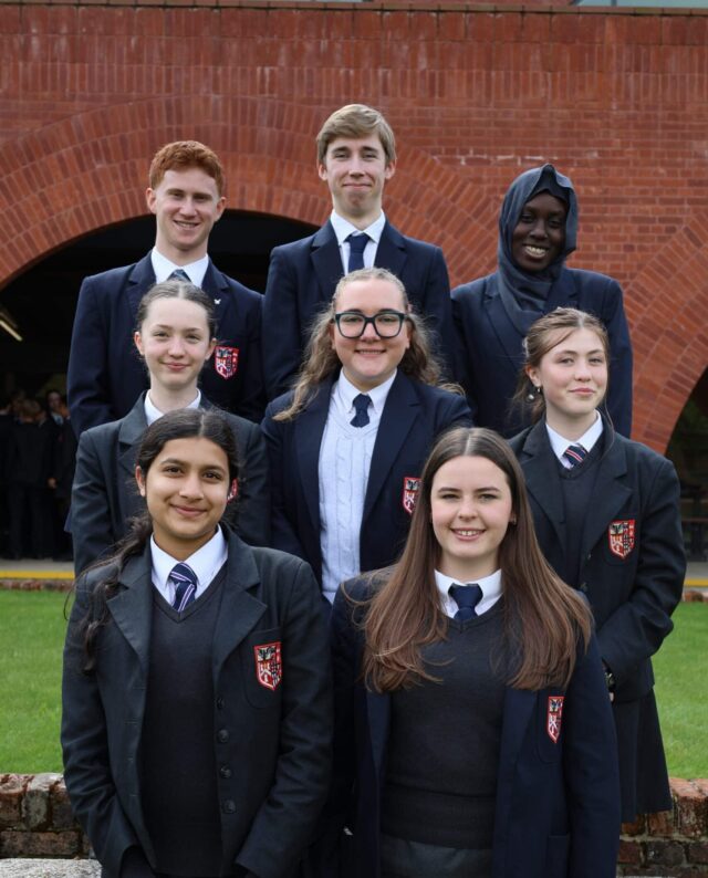 A group of eight students in school uniforms pose outside in front of a brick building, smiling at the camera. Four students stand in the back row, and four in the front row.