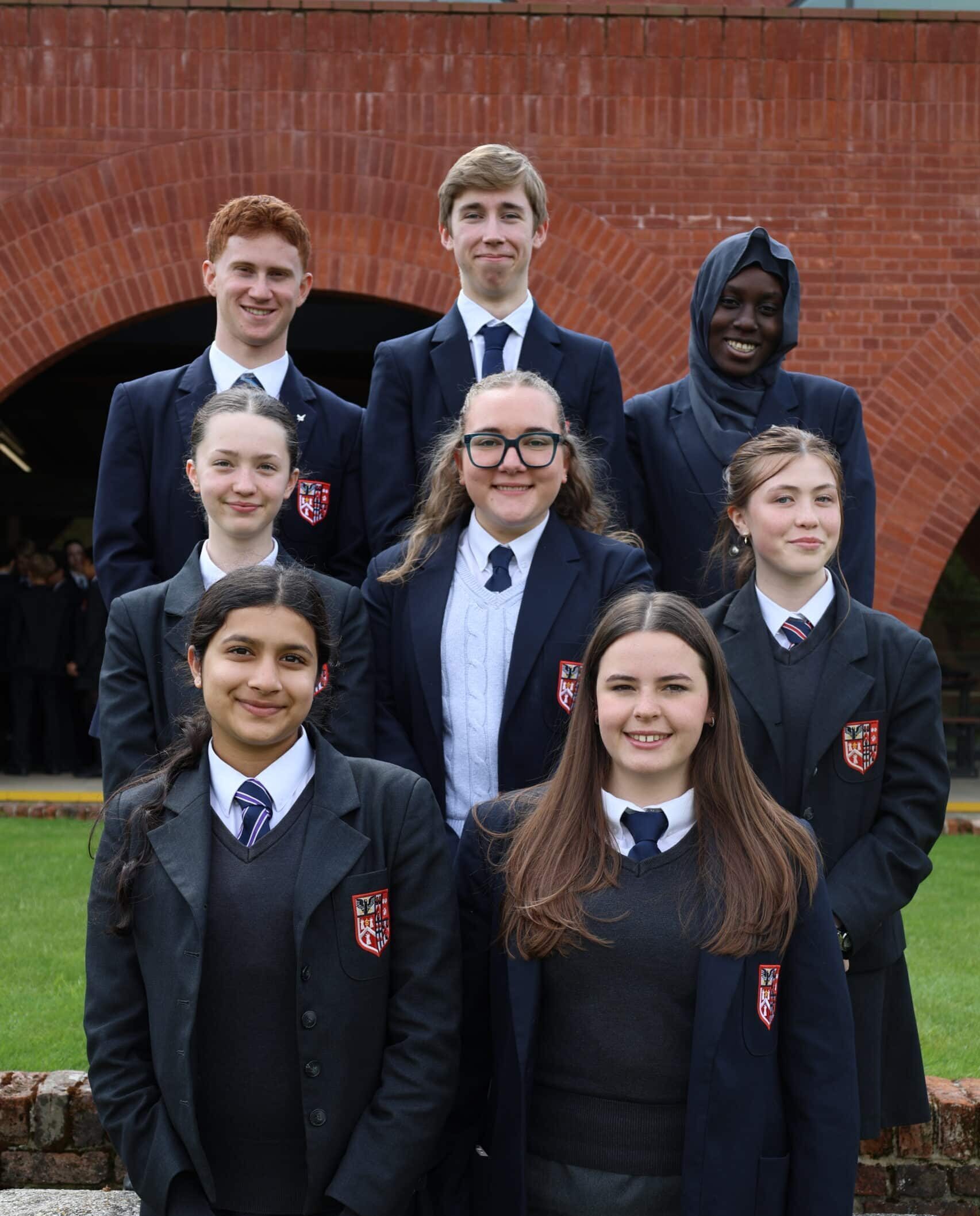 A group of eight students in school uniforms pose outside in front of a brick building, smiling at the camera. Four students stand in the back row, and four in the front row.