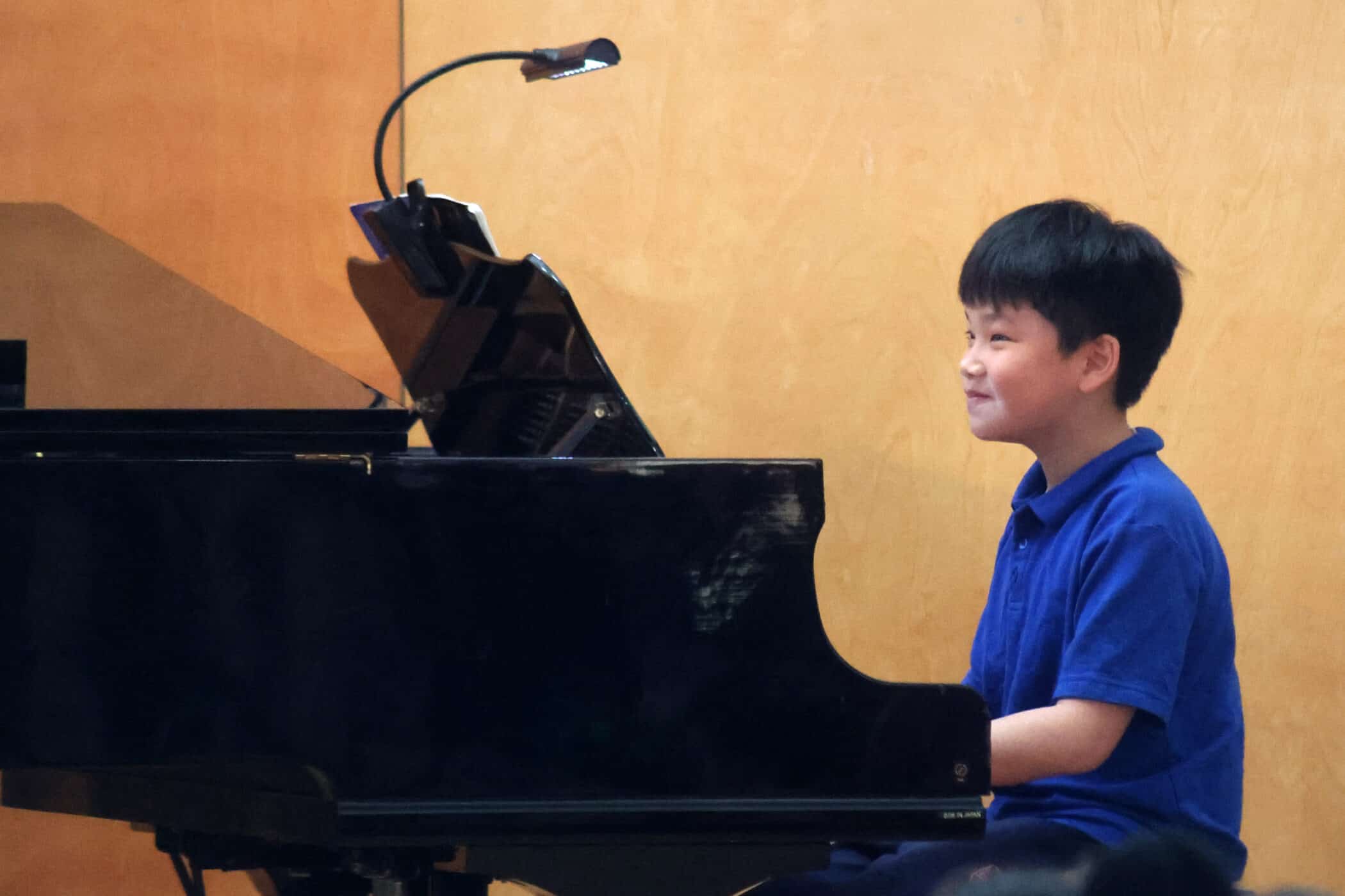 A young boy in a blue shirt sits at a grand piano, smiling as he prepares to play. The background is a plain, light-coloured wall, and sheet music rests on the piano.