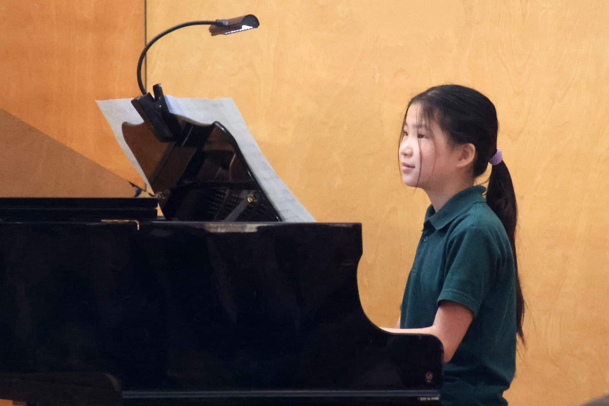 A young girl with a ponytail sits at a black grand piano, looking at sheet music under a small lamp, against a plain yellowish wall. She appears focused and is wearing a green top.