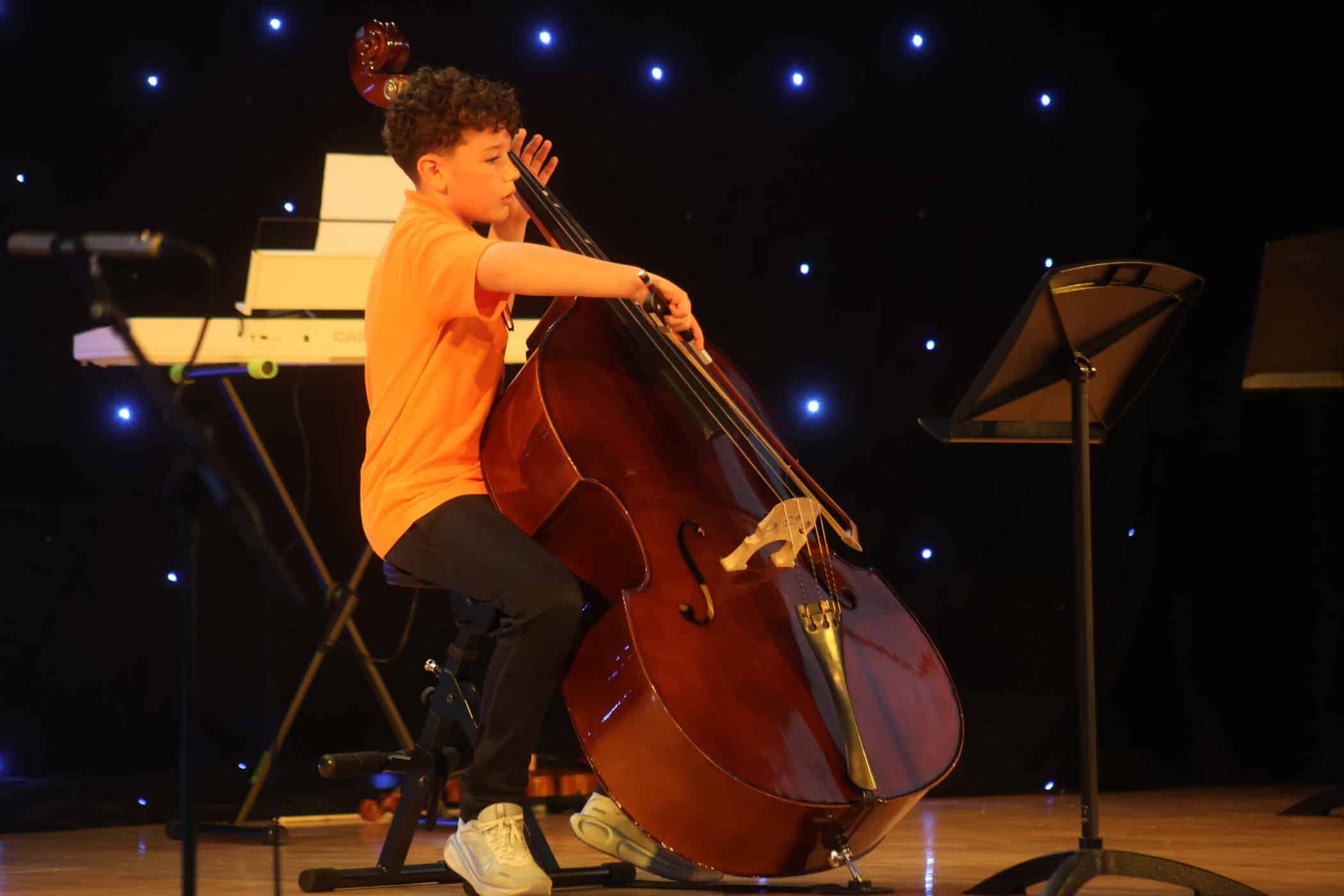 A boy in an orange shirt plays a double bass on stage, with a keyboard and music stand nearby, and a backdrop of small blue lights behind him.