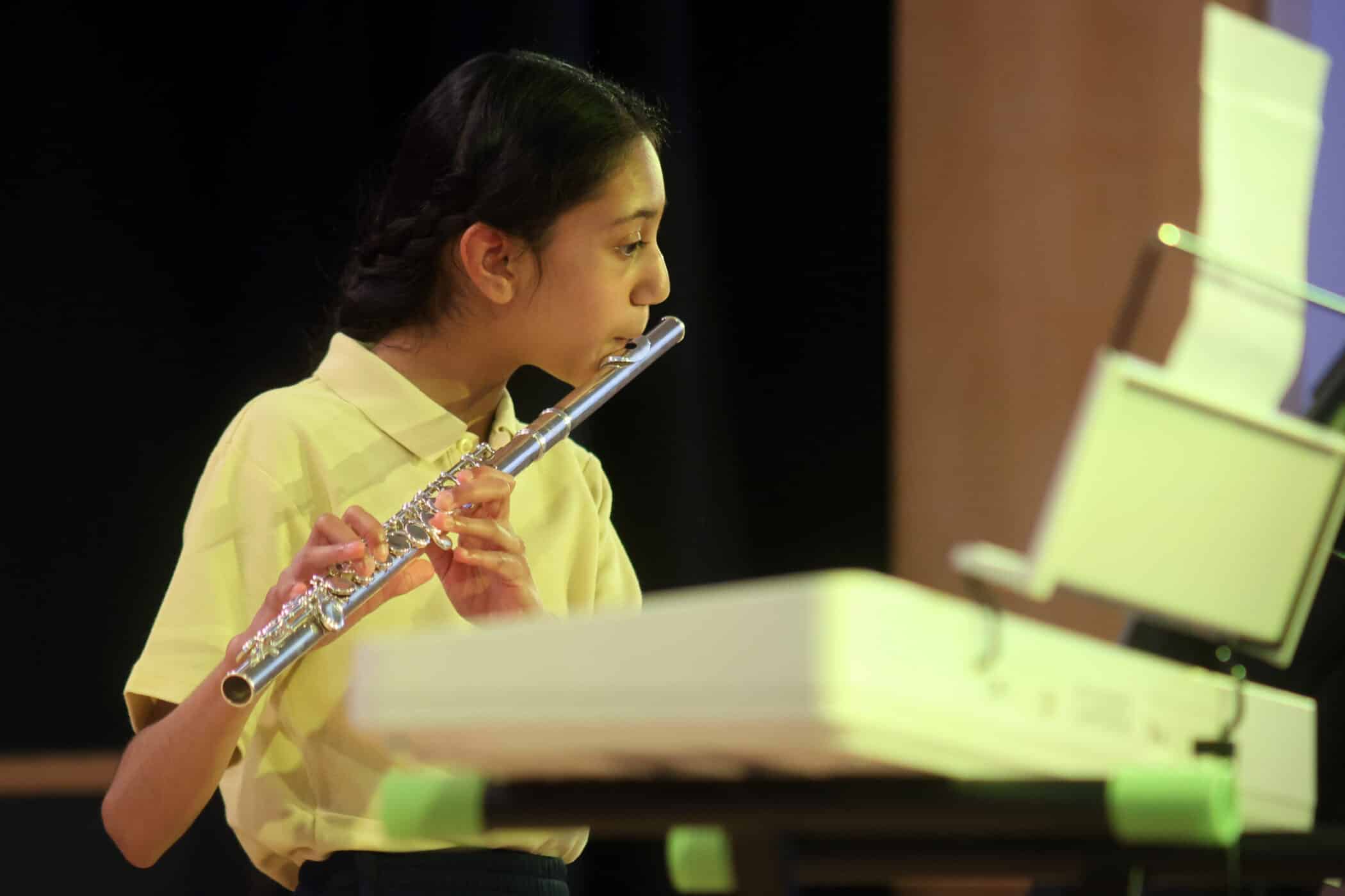 A young girl in a yellow shirt plays the flute whilst reading sheet music on a stand, with a keyboard in front of her and a dark background behind.