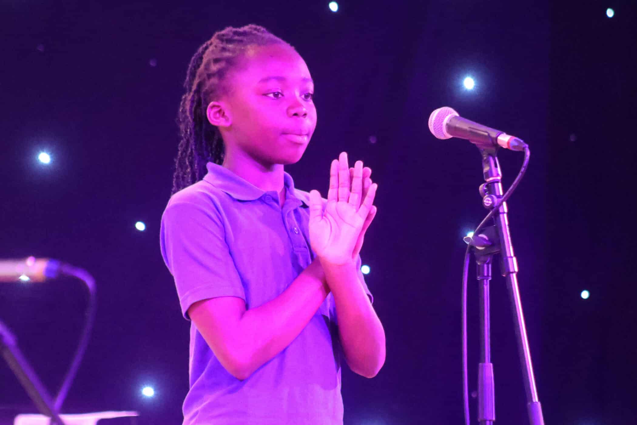 A young person in a purple shirt stands on stage in front of a microphone, holding their hands together. The background is dark with small, bright lights.