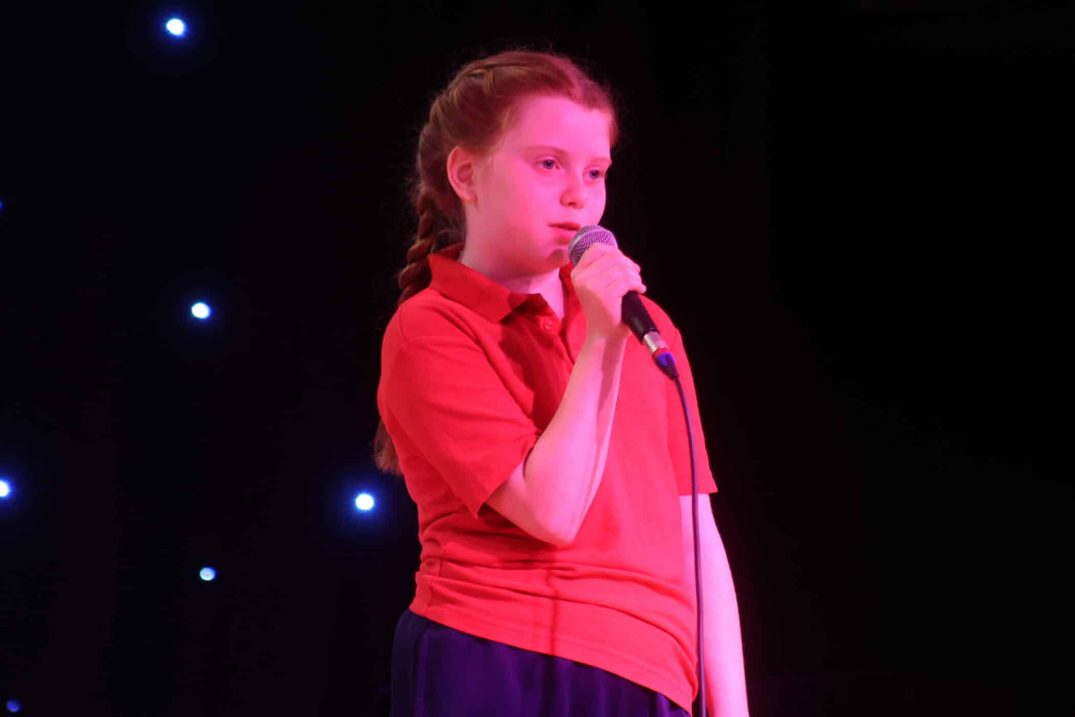 A young girl with red hair in a plait, wearing a red shirt, holds a microphone and stands on stage with small bright lights in the dark background.