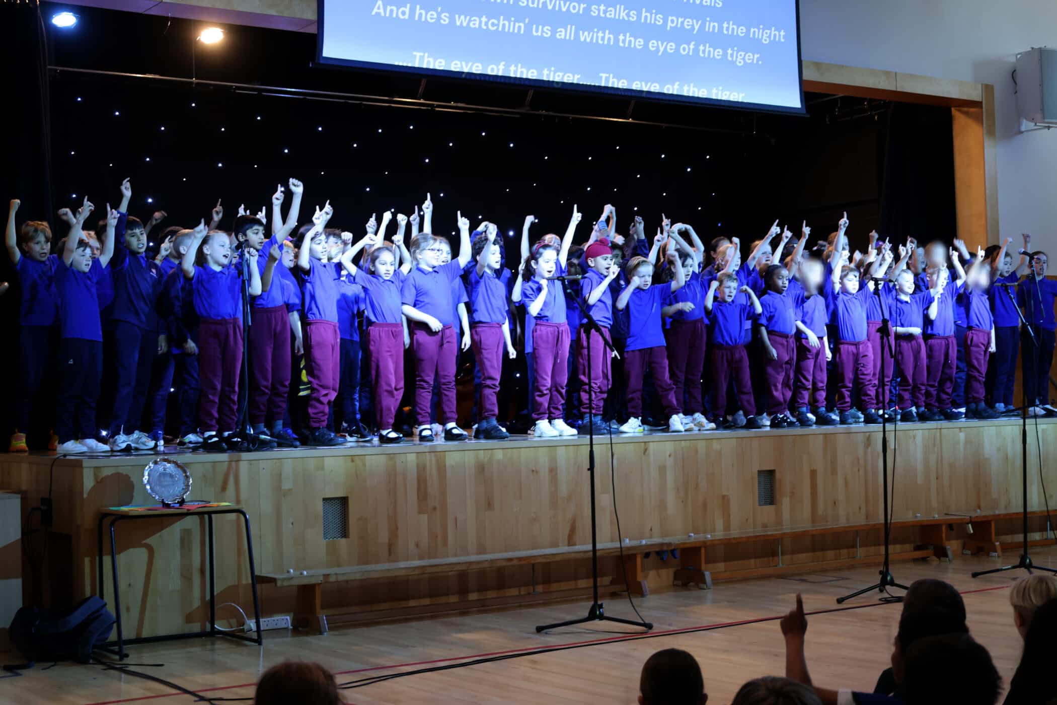 A large group of children in matching purple shirts and maroon trousers stand on a stage, raising their arms in unison during a performance. There are spotlights above and a song’s lyrics projected on a screen behind them.
