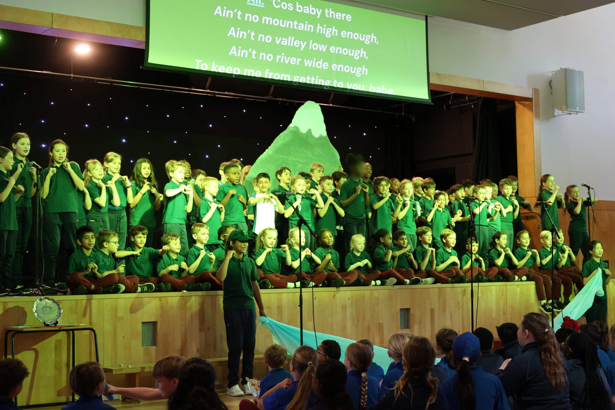 A large group of children in green shirts perform on stage, singing with hand movements in front of a mountain backdrop. Lyrics are projected above them. An audience of children sits and watches.