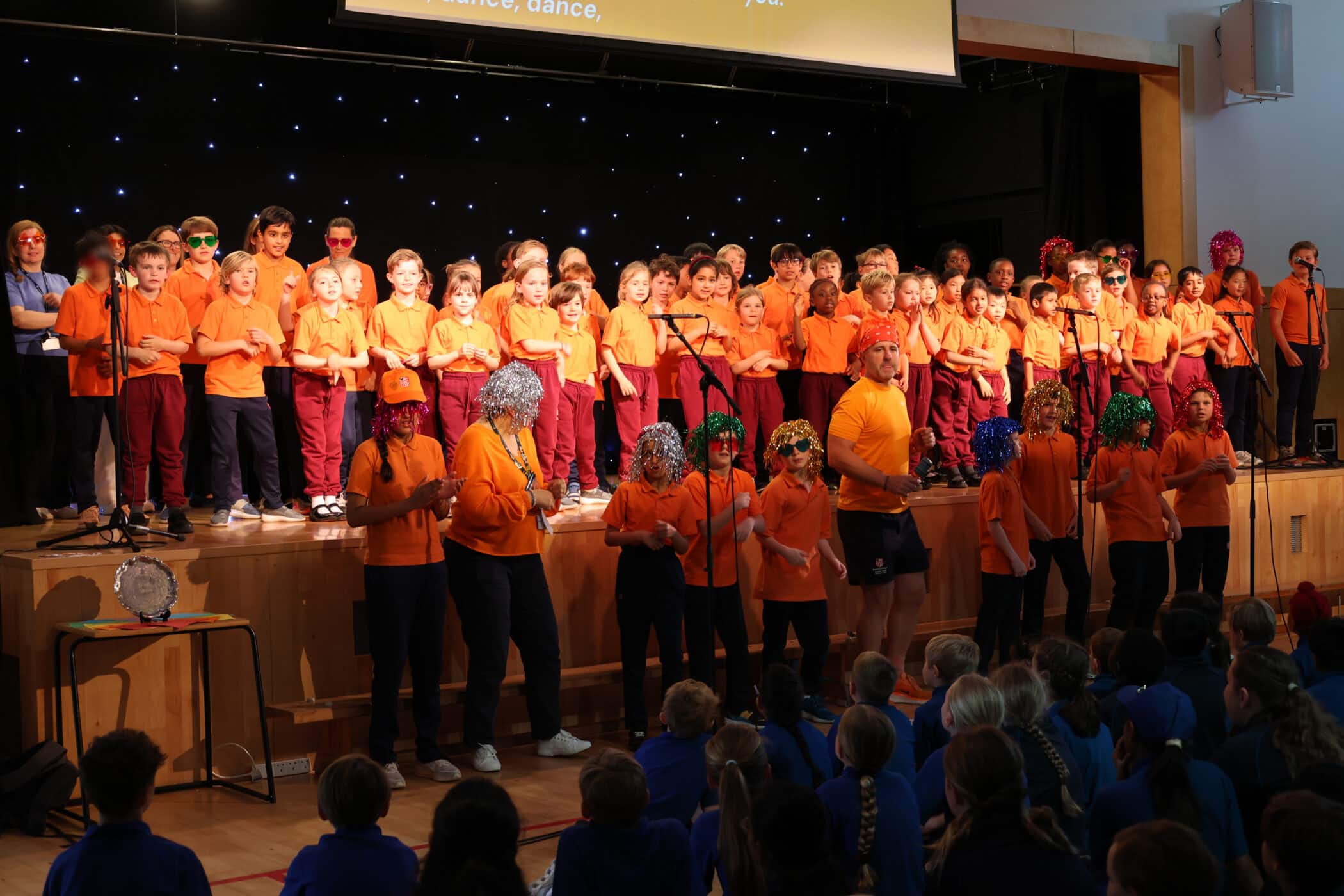 A group of children in orange shirts sing on a stage, with a few adults in front wearing wigs and orange shirts. Seated pupils in blue uniforms watch from the audience in a school hall.