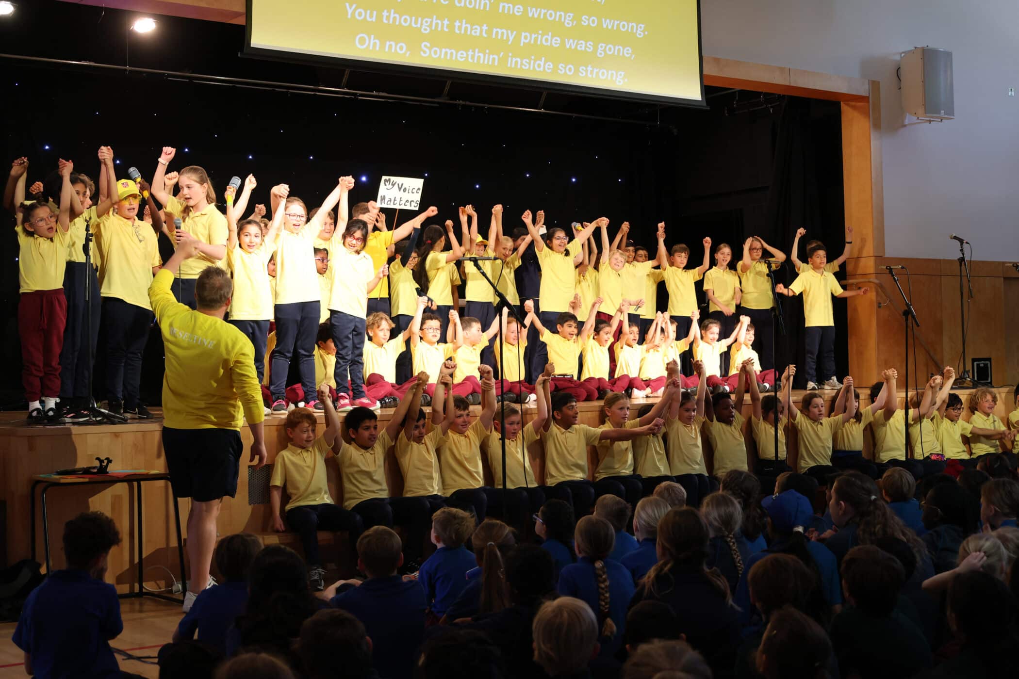 A large group of children in yellow shirts stand on stage with their arms raised, performing for an audience of seated children. A teacher in yellow faces the stage, and song lyrics are projected above.