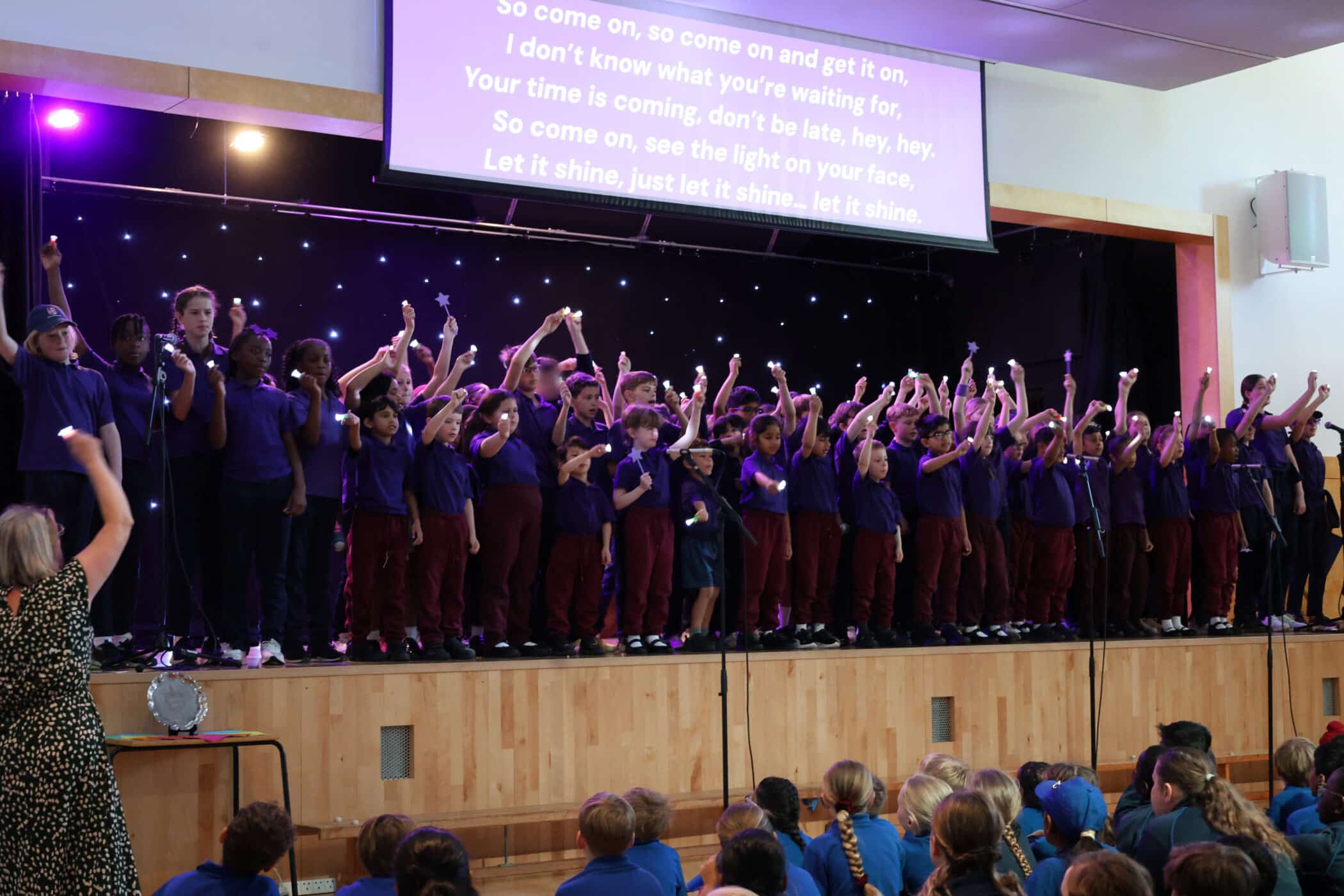 A large group of children in matching purple shirts and maroon trousers perform on stage, raising their arms. An audience sits in front, and song lyrics are projected above them. A teacher stands at the front, leading the performance.