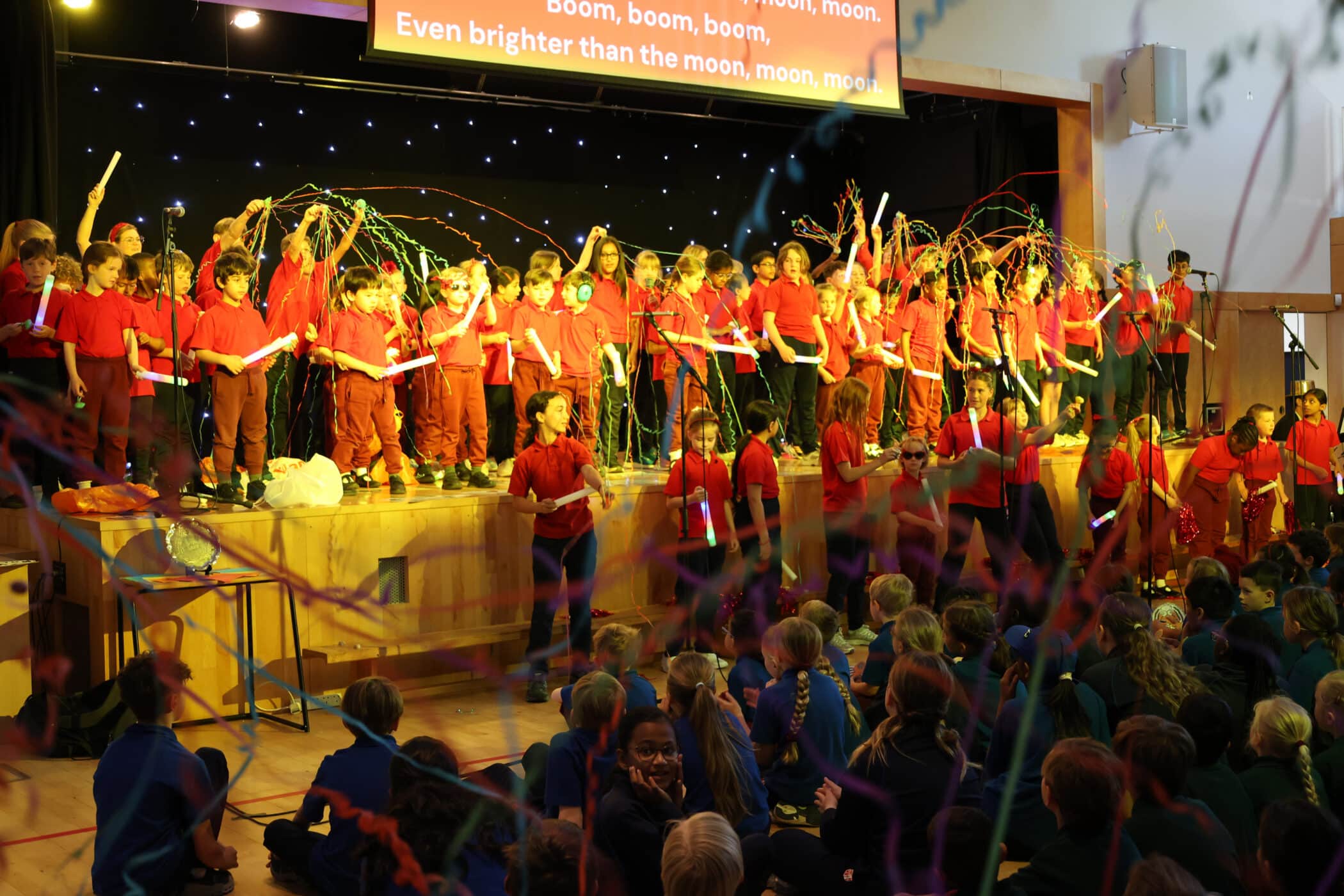 A group of children in red shirts perform on stage holding colourful streamers, while an audience of children in blue and green uniforms watch. Song lyrics are projected above the stage.