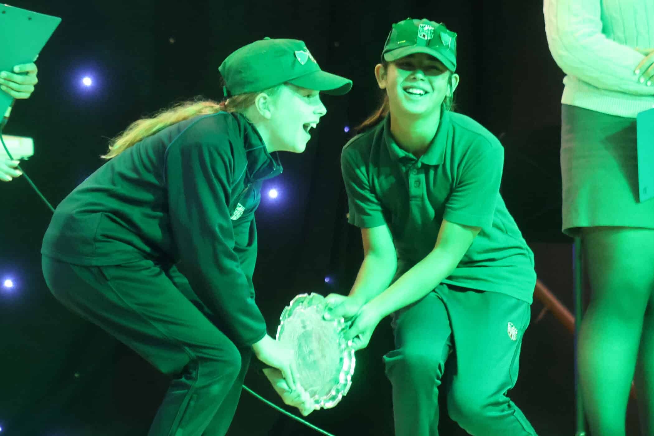 Two children wearing uniforms and caps smile joyfully as they hold a silver trophy plate together on stage, with green lighting and a person standing nearby holding a clipboard.