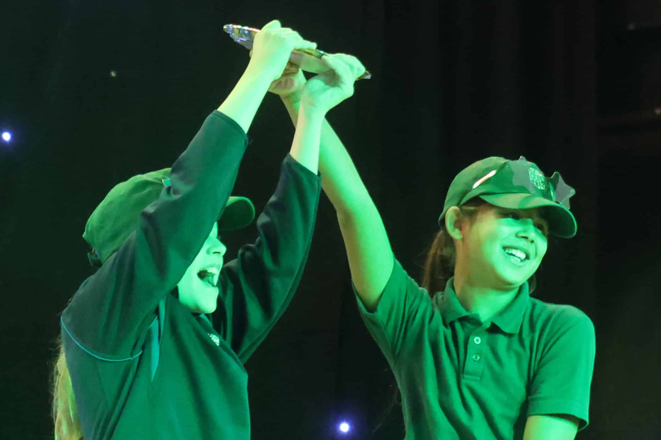 Two children in green uniforms and caps are smiling and holding up a trophy together on stage, celebrating their achievement under bright stage lights.