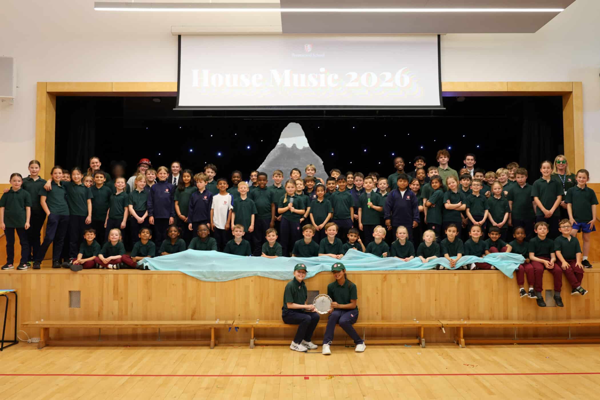 A large group of children and a few adults pose on a stage in a school hall, with a mountain backdrop and a sign that reads House Music 2026. Two pupils sit front and centre holding a trophy.