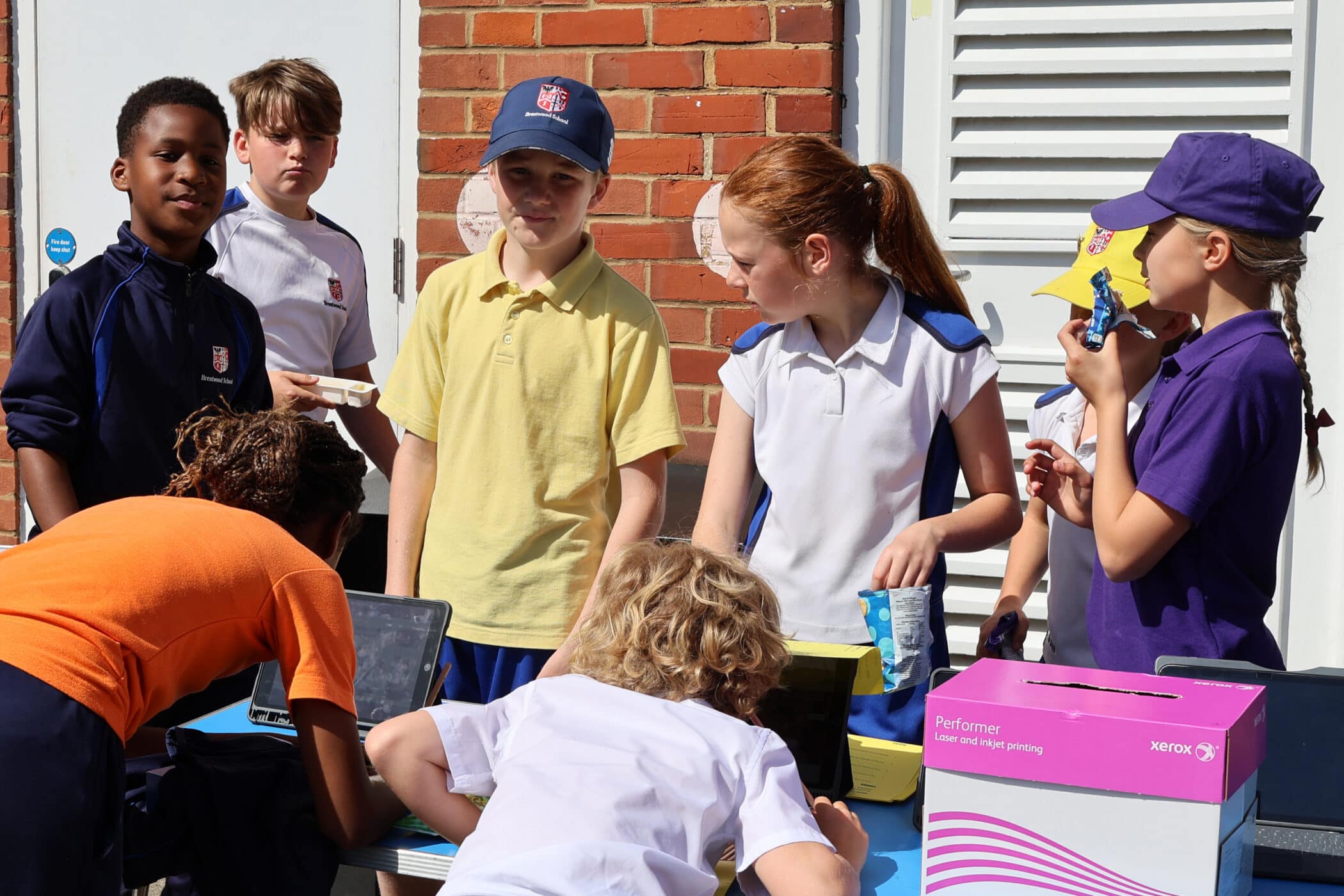 A group of children wearing sports kits gather around a table outdoors, some using laptops and others talking or eating snacks, with a brick wall and boxes visible in the background.