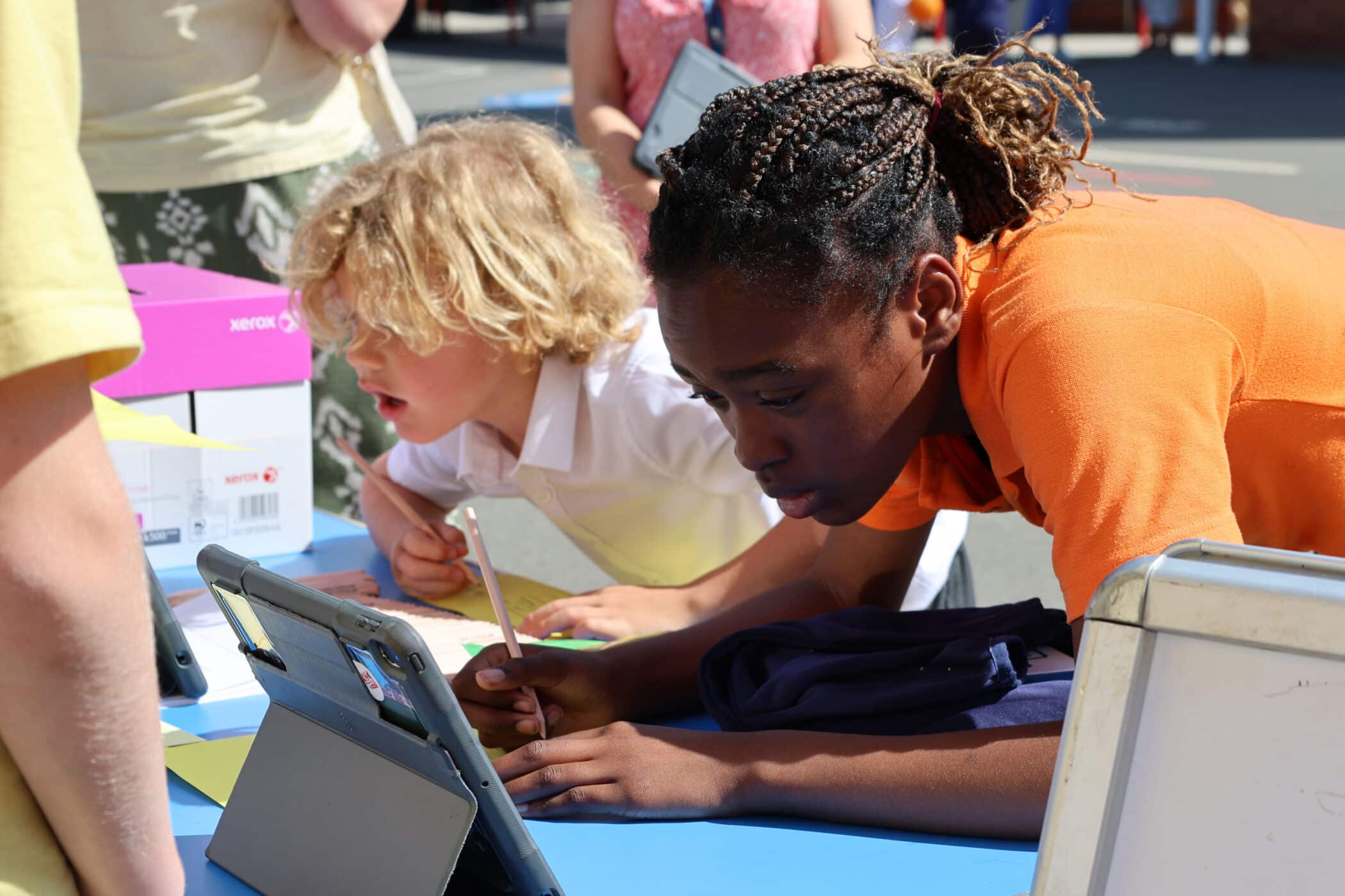 Two children lean over a blue table, focused on writing or drawing on paper and using tablets. The girl in an orange shirt writes intently, whilst the boy beside her, with blond curly hair, looks on. Other people are nearby.