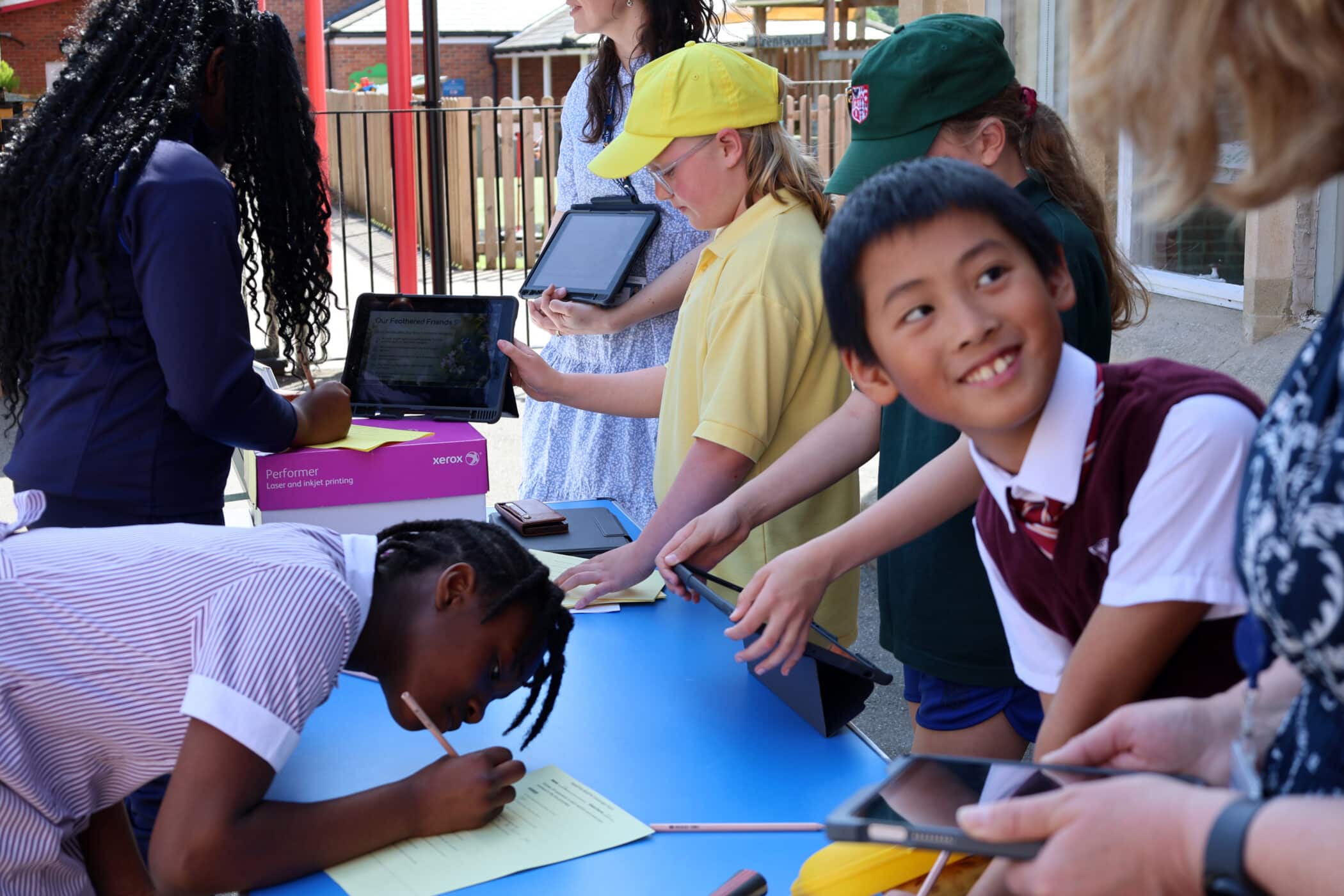 Children in school uniforms use tablets and fill out papers at a blue table outdoors, supervised by adults. Some are standing whilst one girl leans over to write, and everyone is engaged in their activities.