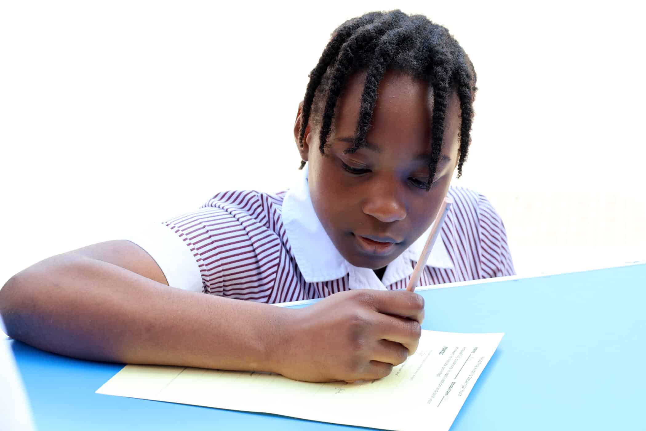 A young girl in a striped school uniform writes on a sheet of paper at a blue desk, concentrating on her work.