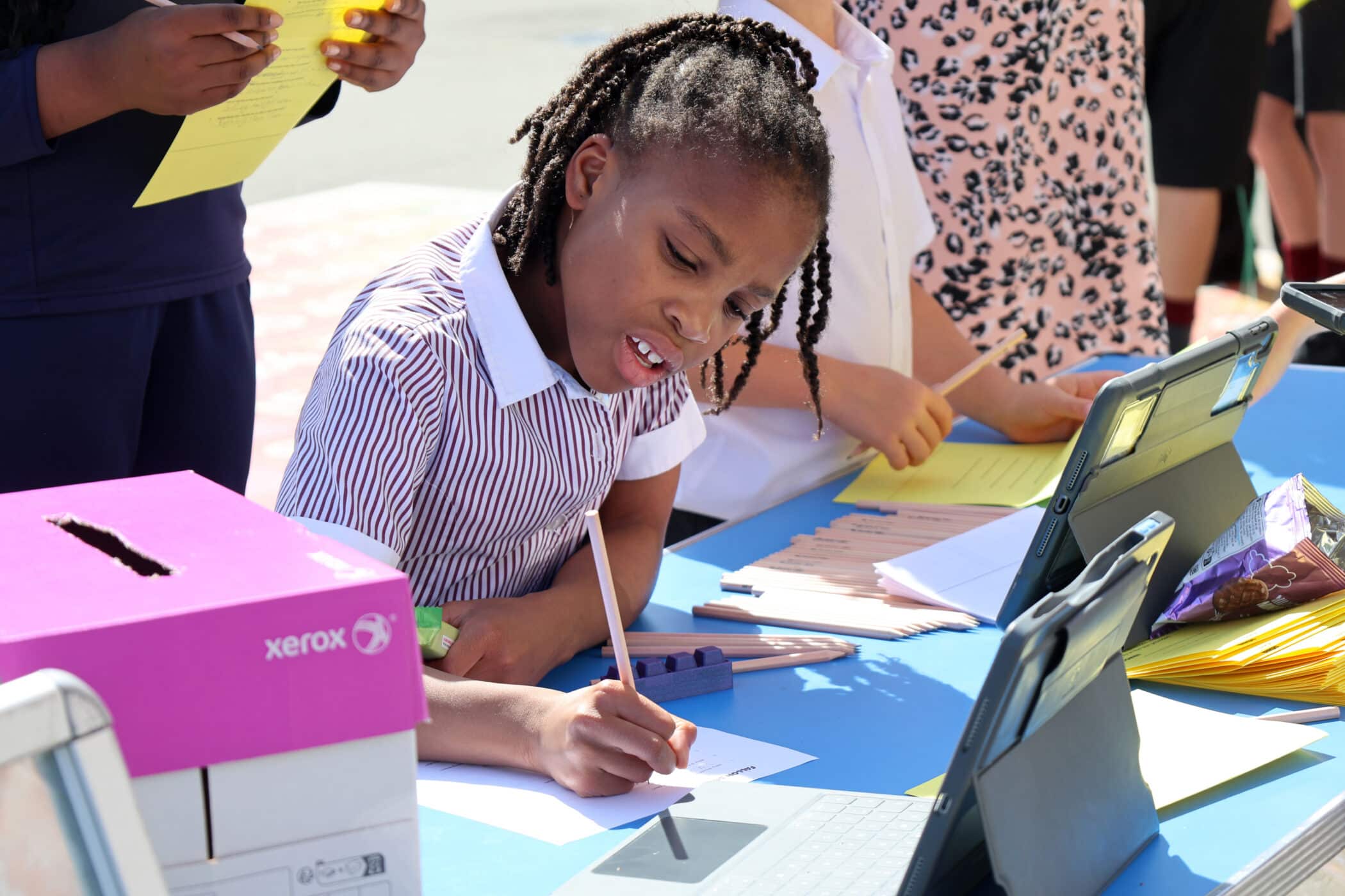 A young girl with plaits writes on paper at an outdoor table with tablets, papers, and school supplies. Other children are around her, holding yellow sheets. It appears to be a school or community event on a sunny day.