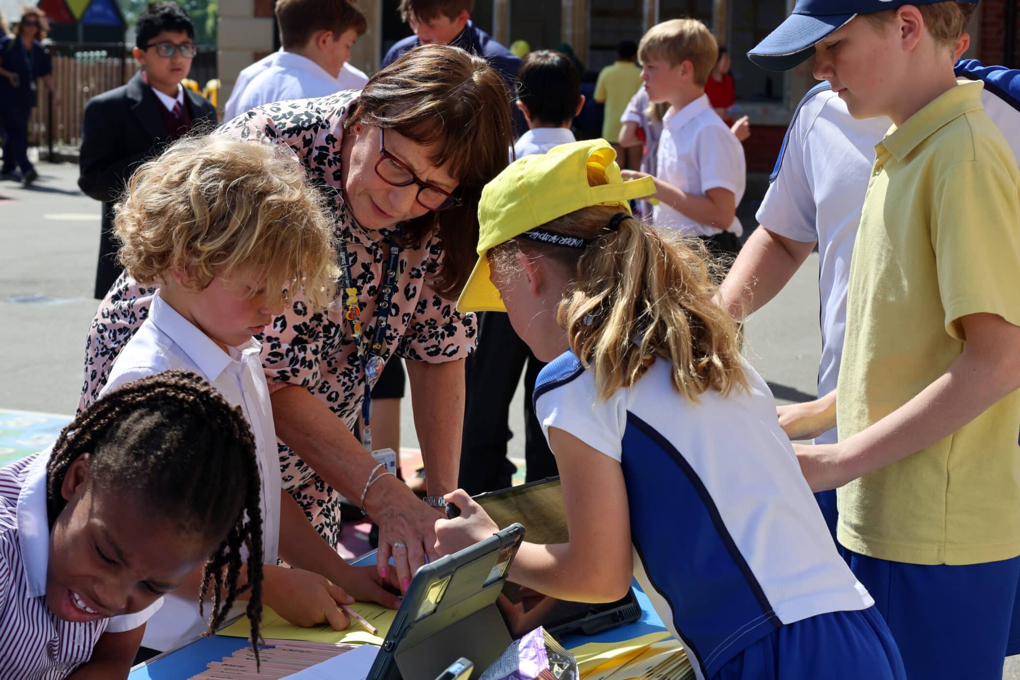 A teacher helps a group of young pupils with an outdoor activity at a table, whilst other children play and interact in the background on a sunny day.