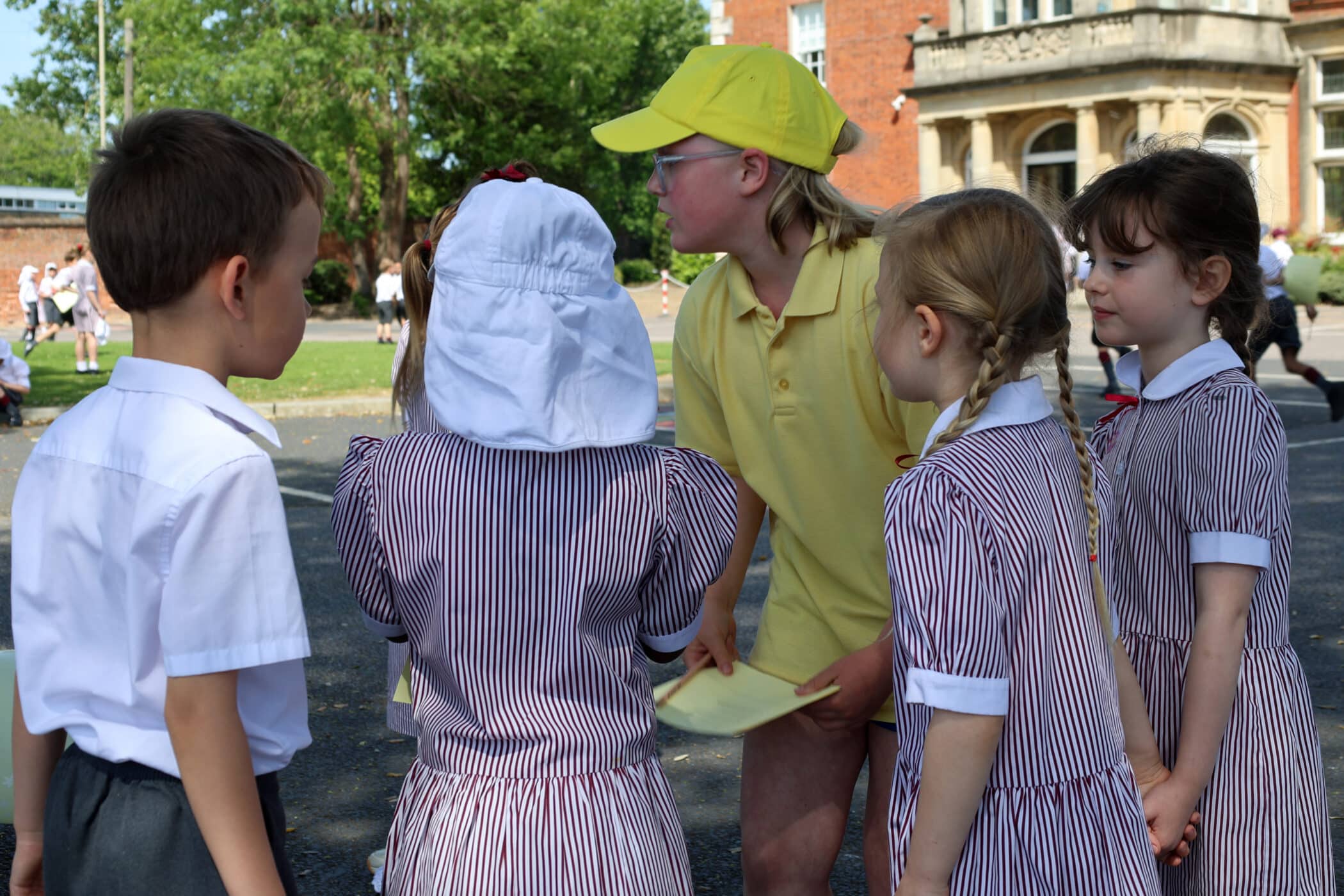 A group of young children in school uniforms stand together outside. One child in a yellow cap and shirt appears to be talking to the others, who are wearing striped uniforms. Trees and a building are in the background.