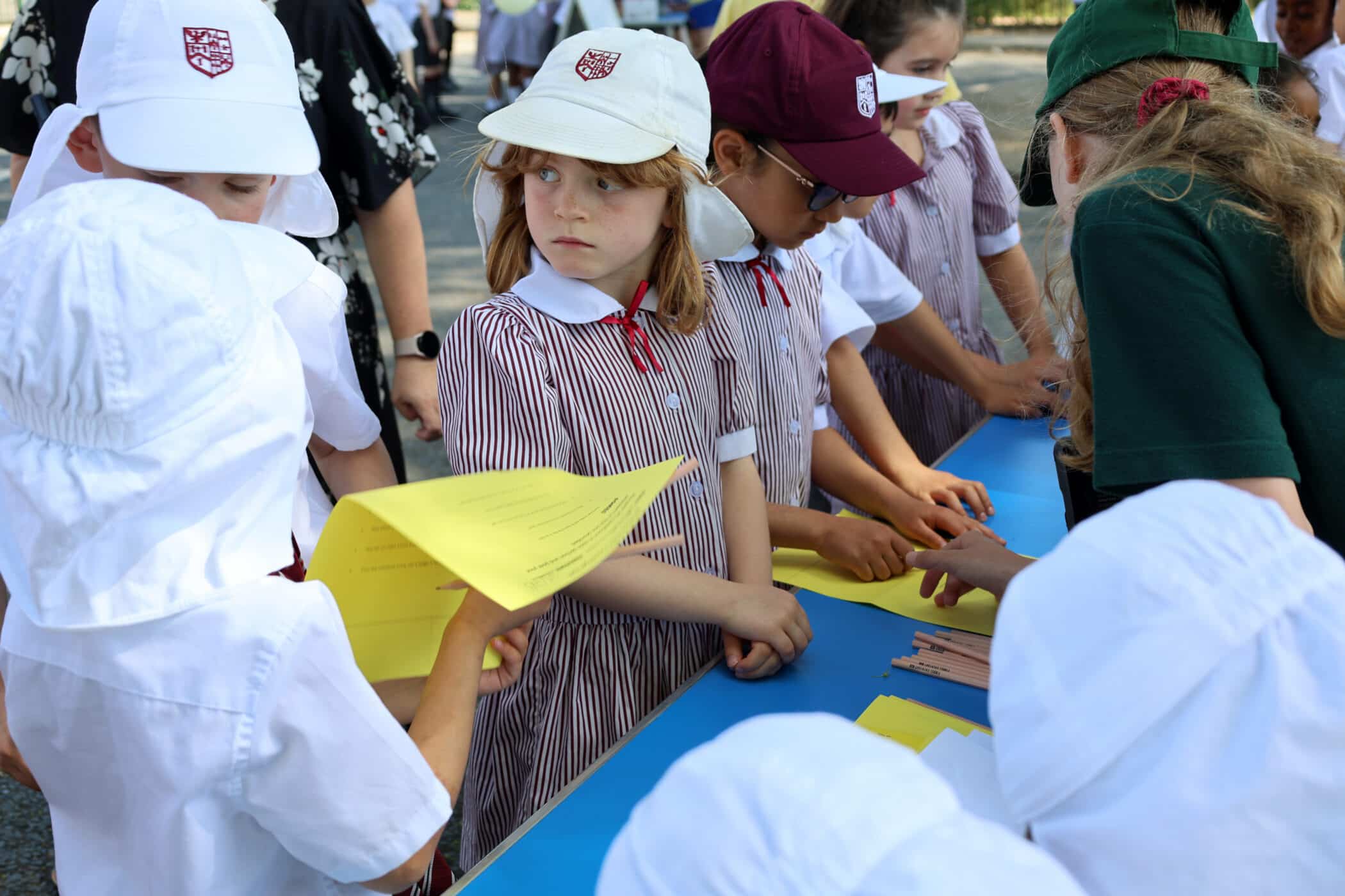 A group of school children in uniforms and hats gather round a blue table outdoors, holding yellow papers and interacting with an adult wearing a green shirt.