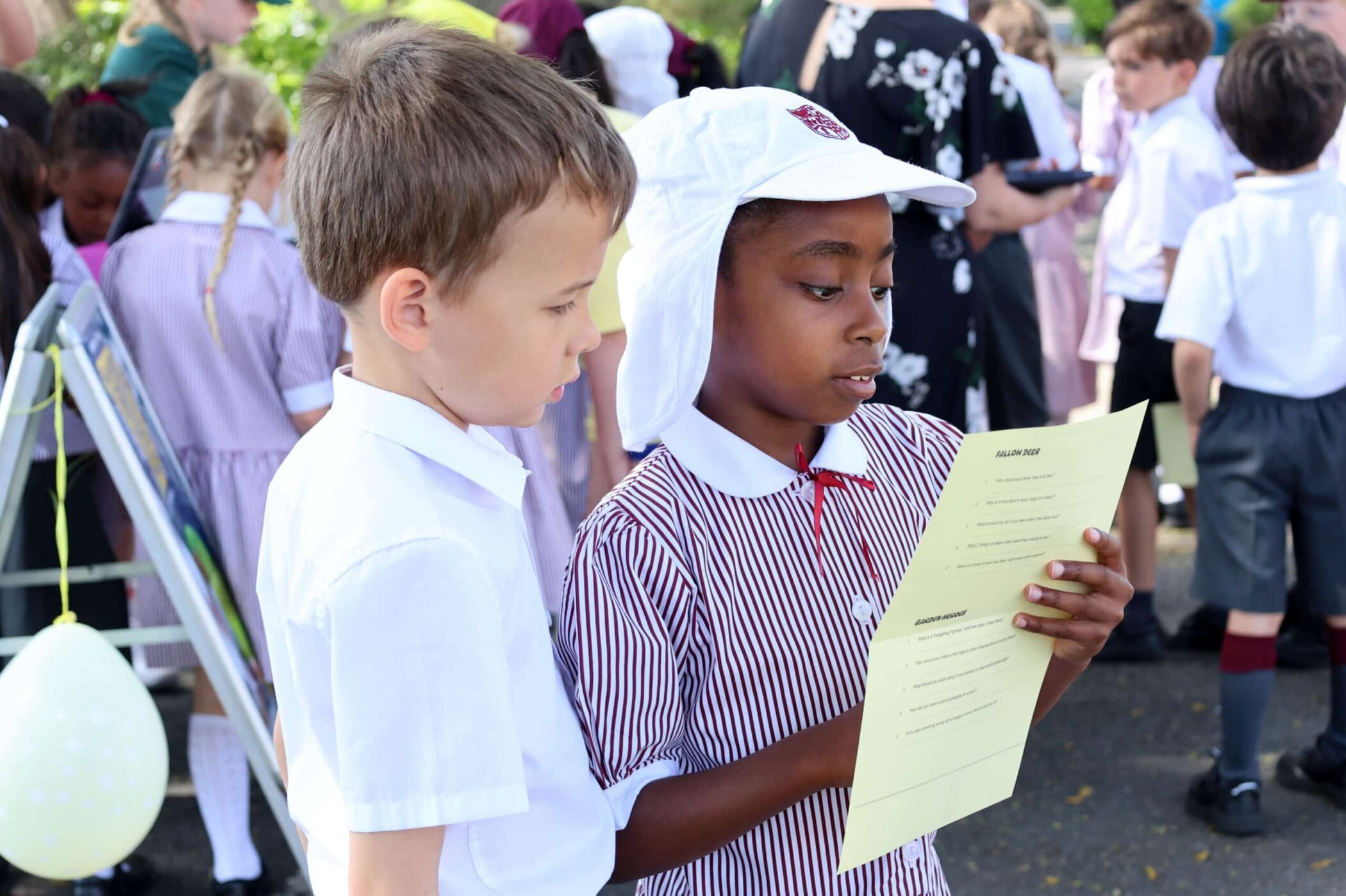 Two schoolchildren, one in a white shirt and one in a striped dress and white hat, stand closely together outdoors, reading a yellow paper. Other children in uniforms are gathered in the background.
