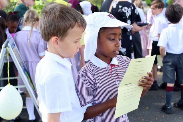 Two schoolchildren, one in a white shirt and one in a striped dress and white hat, stand closely together outdoors, reading a yellow paper. Other children in uniforms are gathered in the background.