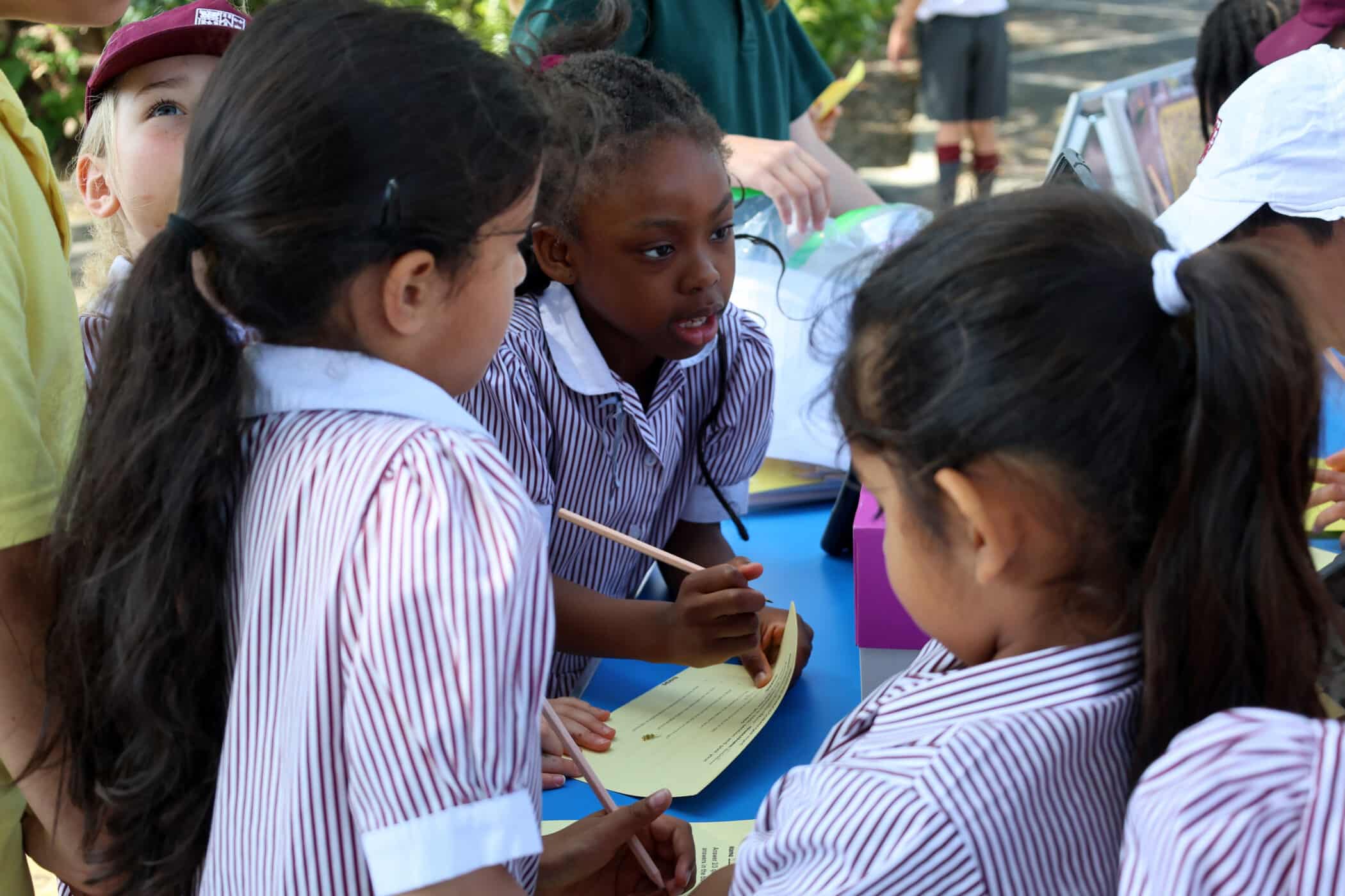 A group of young girls in striped school uniforms gather around a blue table outdoors, working together on an activity with paper and pencils.