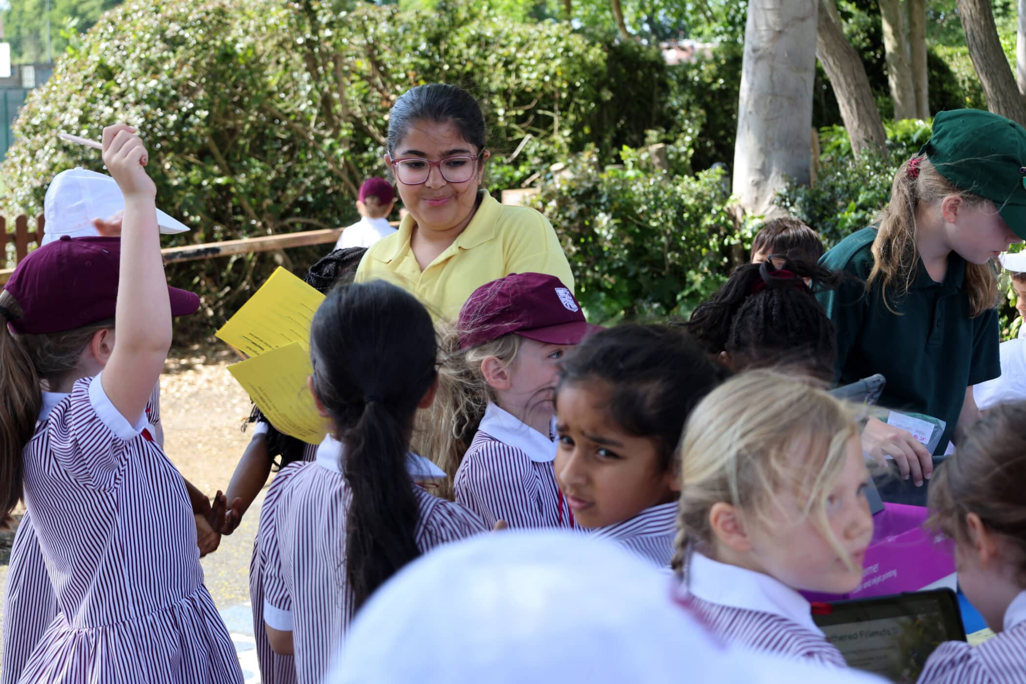 A group of young girls in striped uniforms and hats gather outdoors around a table, with a woman in a yellow shirt holding papers and smiling at them. Trees and greenery are visible in the background.